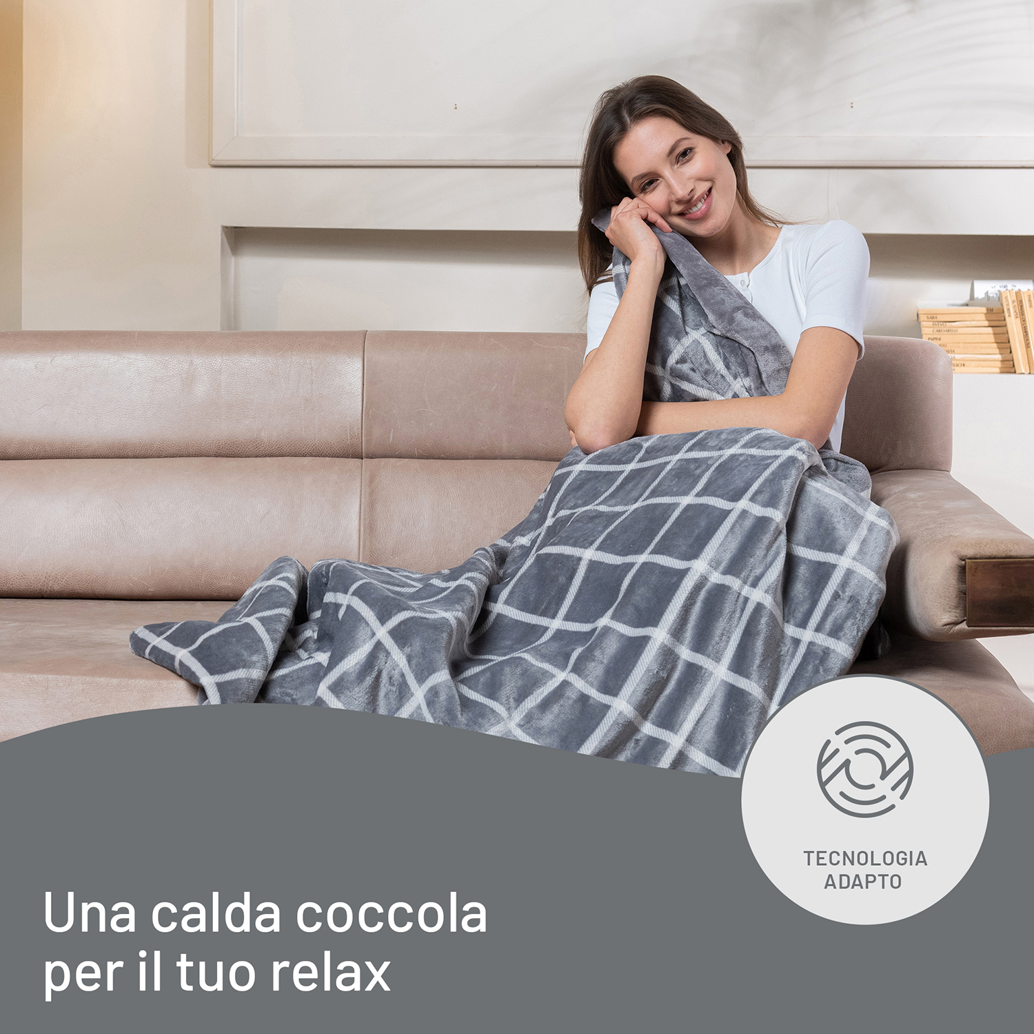 Una donna sorridente, coperta da una coperta grigia, seduta su un divano beige.