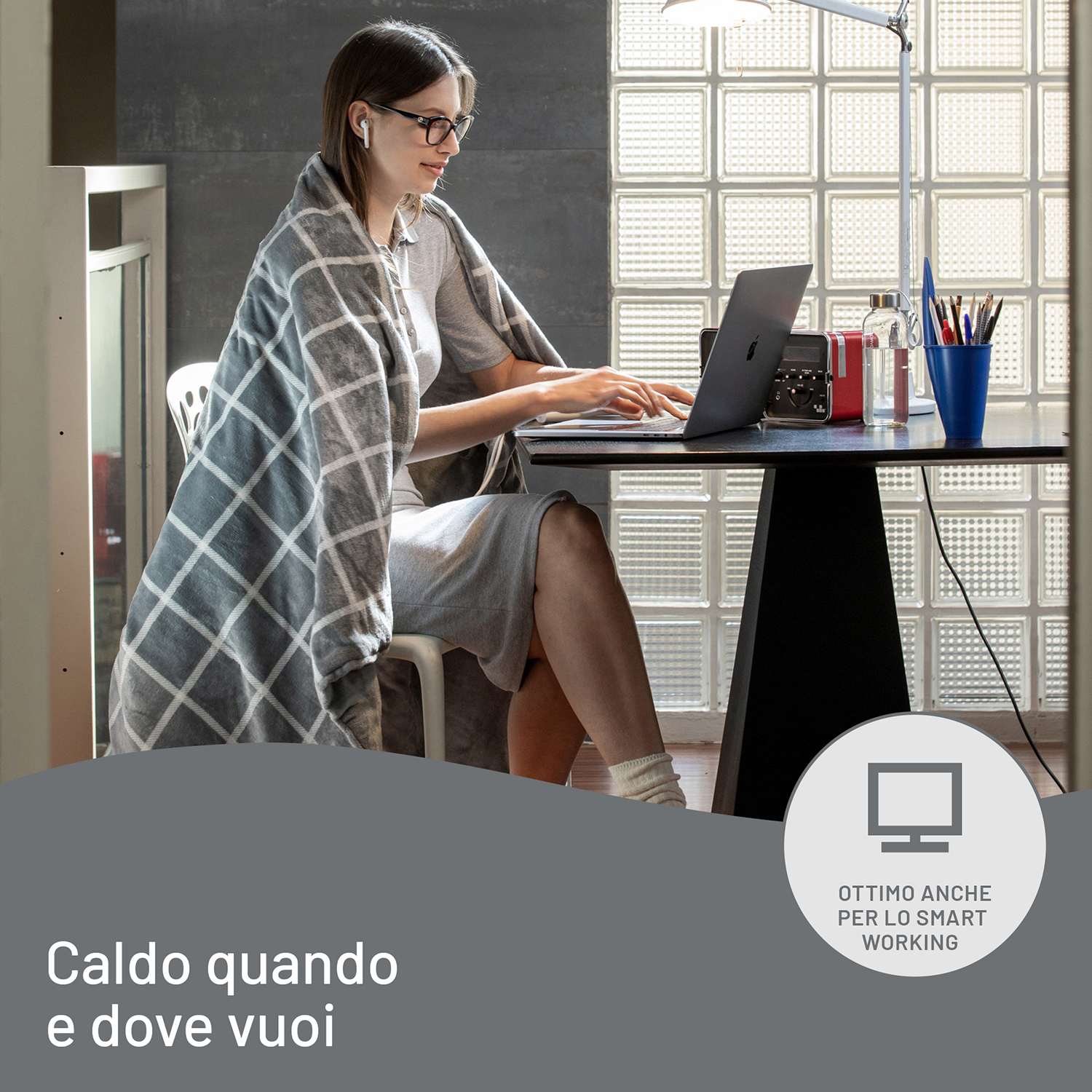 Donna che lavora al computer portatile, con occhiali e coperta in una stanza. Sfondo mattone.