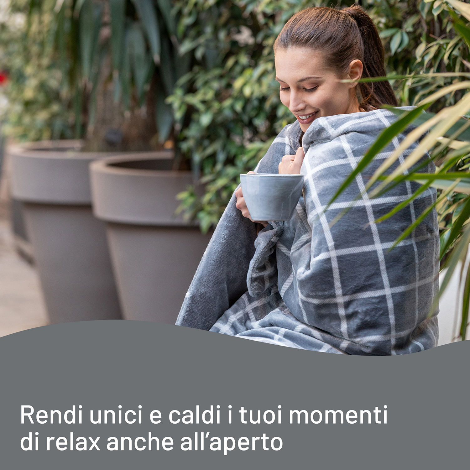 Donna avvolta in coperta a quadri, con tazza, vicino a grandi vasi.