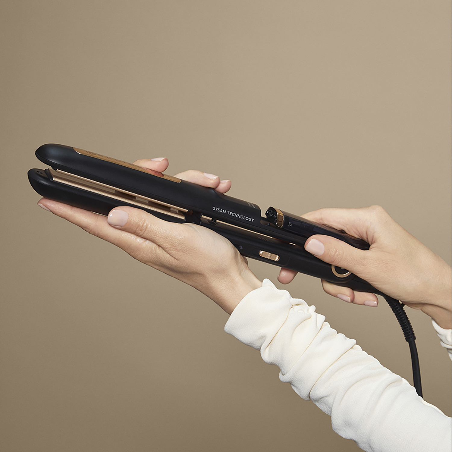 Mani che tengono una piastra per capelli nera e oro. Piastre nere con dettagli oro. Sfondo beige chiaro.
