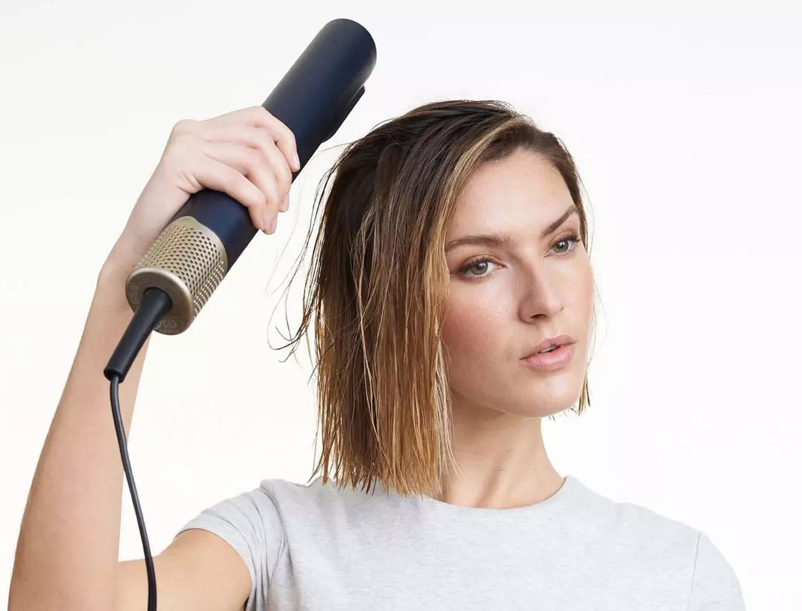 Kobieta trzyma czarno-złotą suszarkę do włosów. Ma mokre włosy.