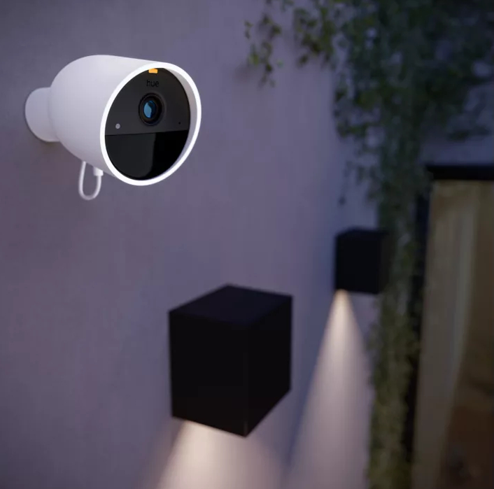 Biała kamera bezpieczeństwa Philips Hue zamontowana na ścianie z dwoma czarnymi światłami.