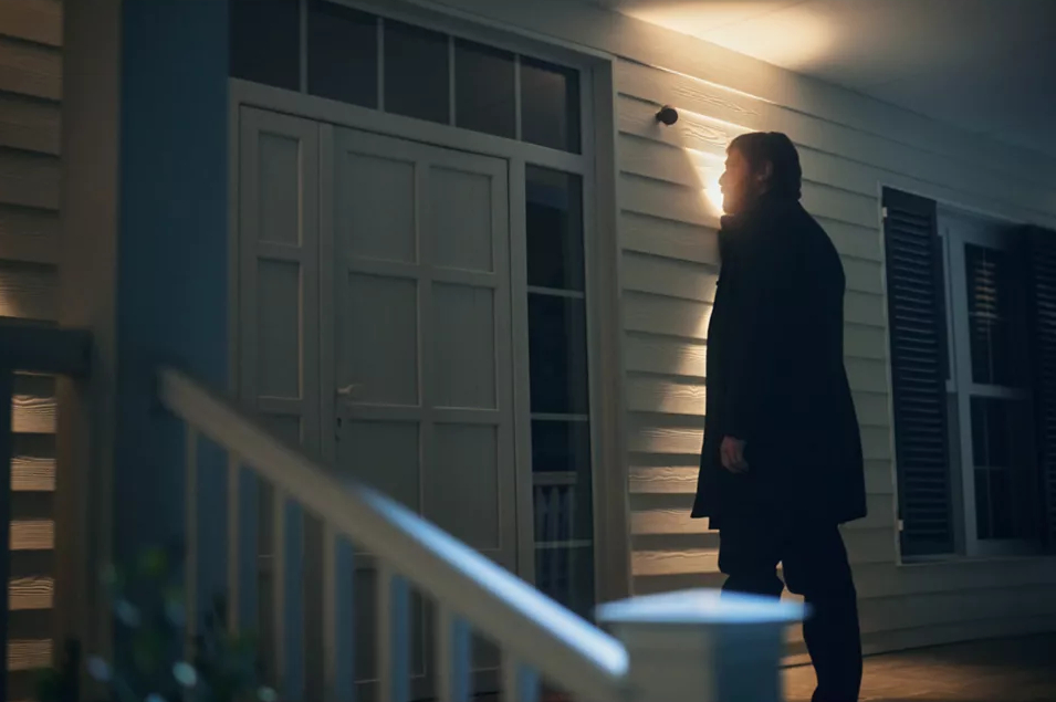 Mężczyzna w ciemnym płaszczu idzie w kierunku domu o zmierzchu, oświetlony.