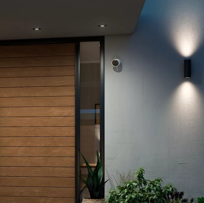 Widok zewnętrzny nowoczesnego wejścia do domu z drewnianymi drzwiami, kamerą i zielonymi roślinami.