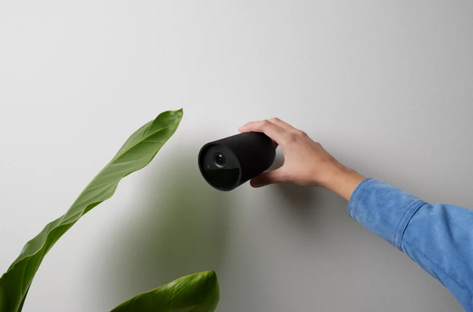 Osoba montująca czarną kamerę bezpieczeństwa na białej ścianie, z zieloną rośliną w pobliżu.