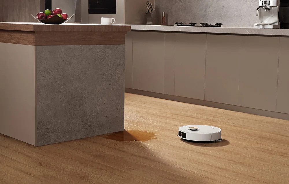 Robot odkurzający czyści drewnianą podłogę. W pobliżu wyspy kuchennej jest rozlana woda.