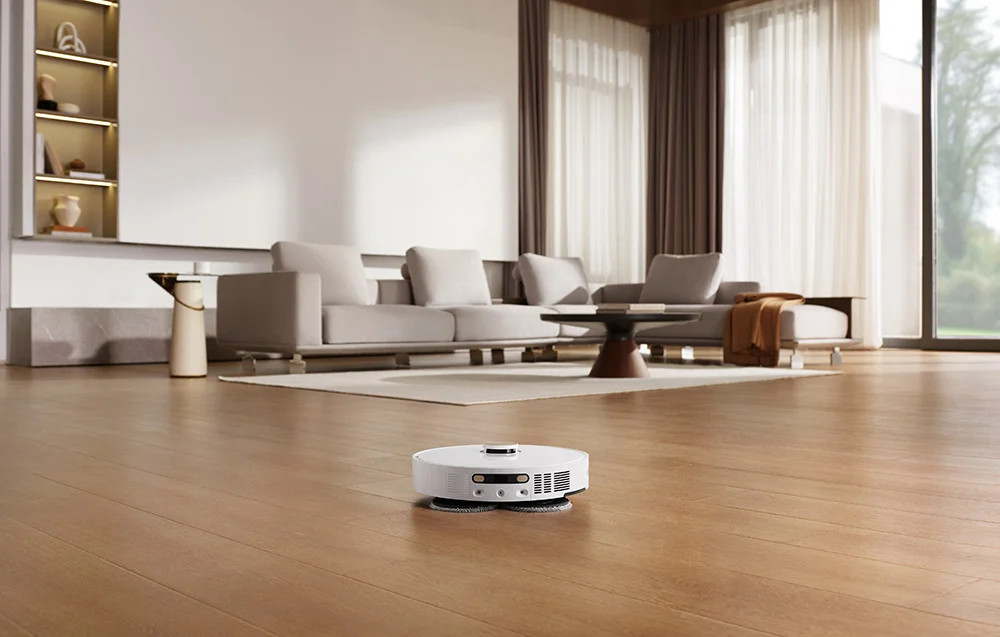 Biały robot sprzątający na drewnianej podłodze. W tle sofa i okno.