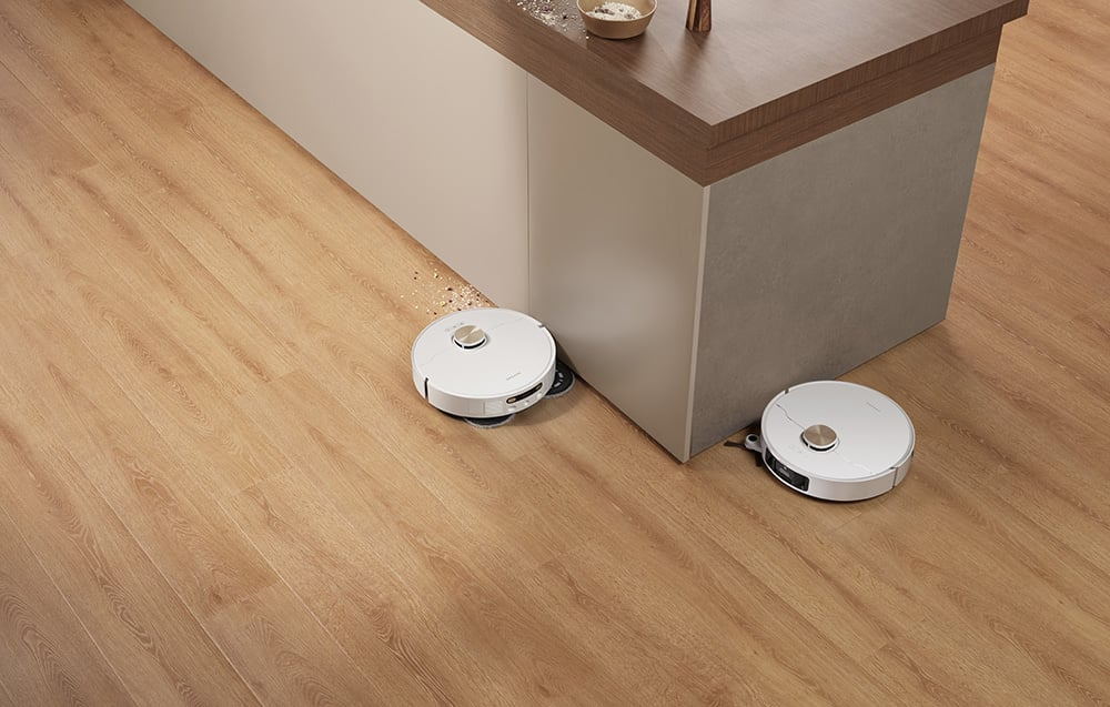 Dwa białe roboty sprzątające na drewnianej podłodze obok białego blatu z drewnianym blatem.