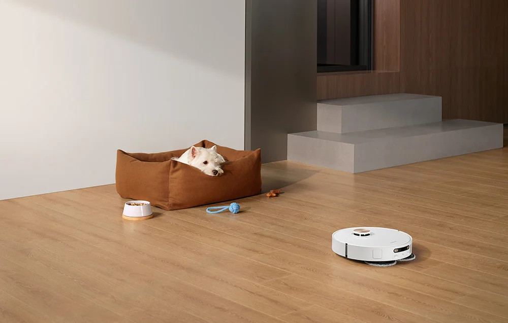 Biały pies w brązowym legowisku i robot odkurzający w pokoju z drewnianą podłogą.