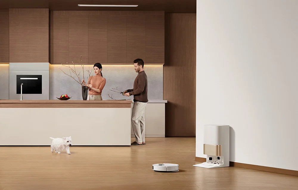 Para aranżuje gałęzie w kuchni, robot sprzątający i pies na podłodze.