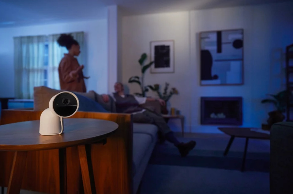 Biała kamera bezpieczeństwa na stole w salonie. Dwie osoby w tle.
