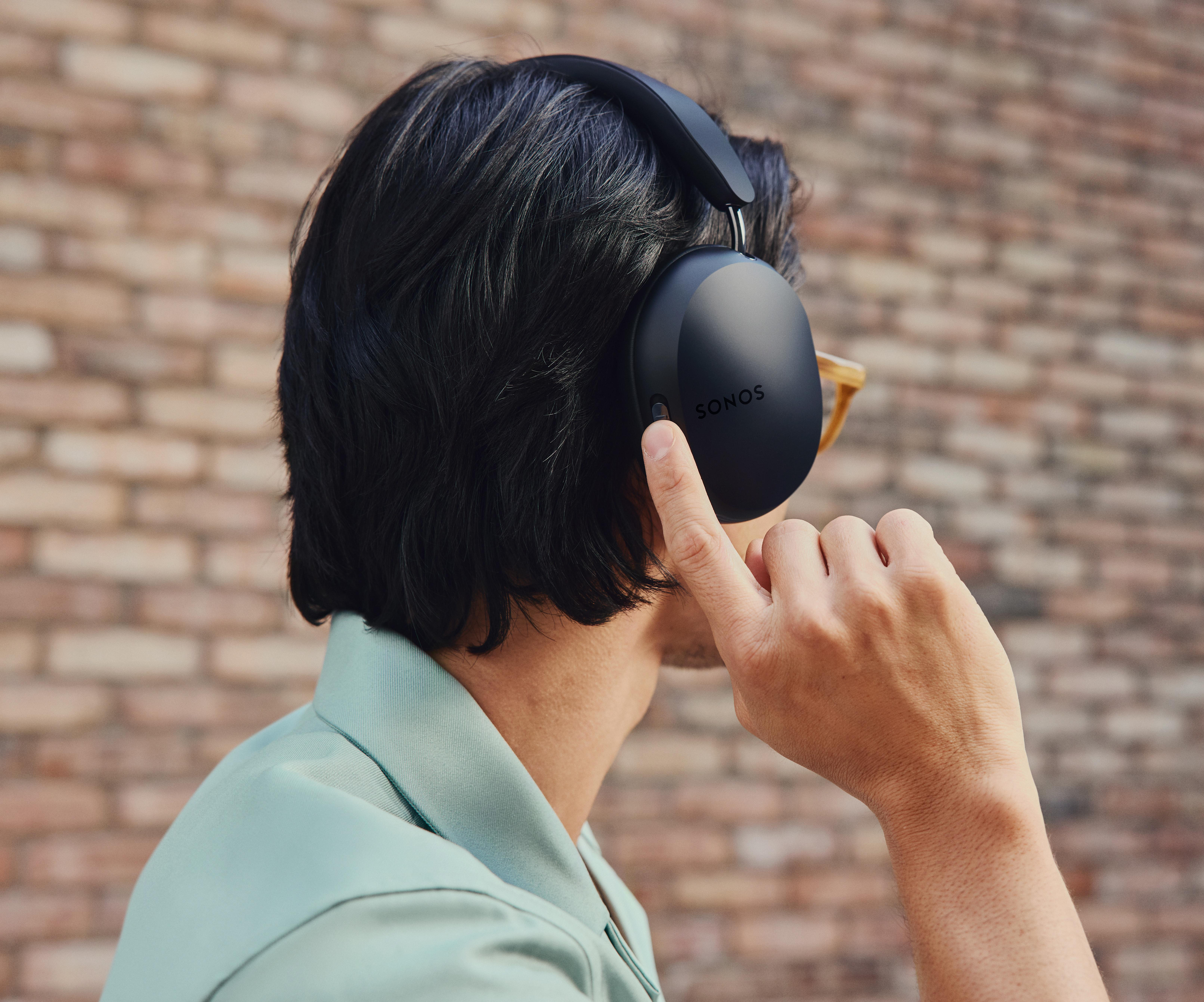 Person trägt schwarze Sonos-Kopfhörer, zeigt auf die rechte Ohrmuschel, Ziegelhintergrund.