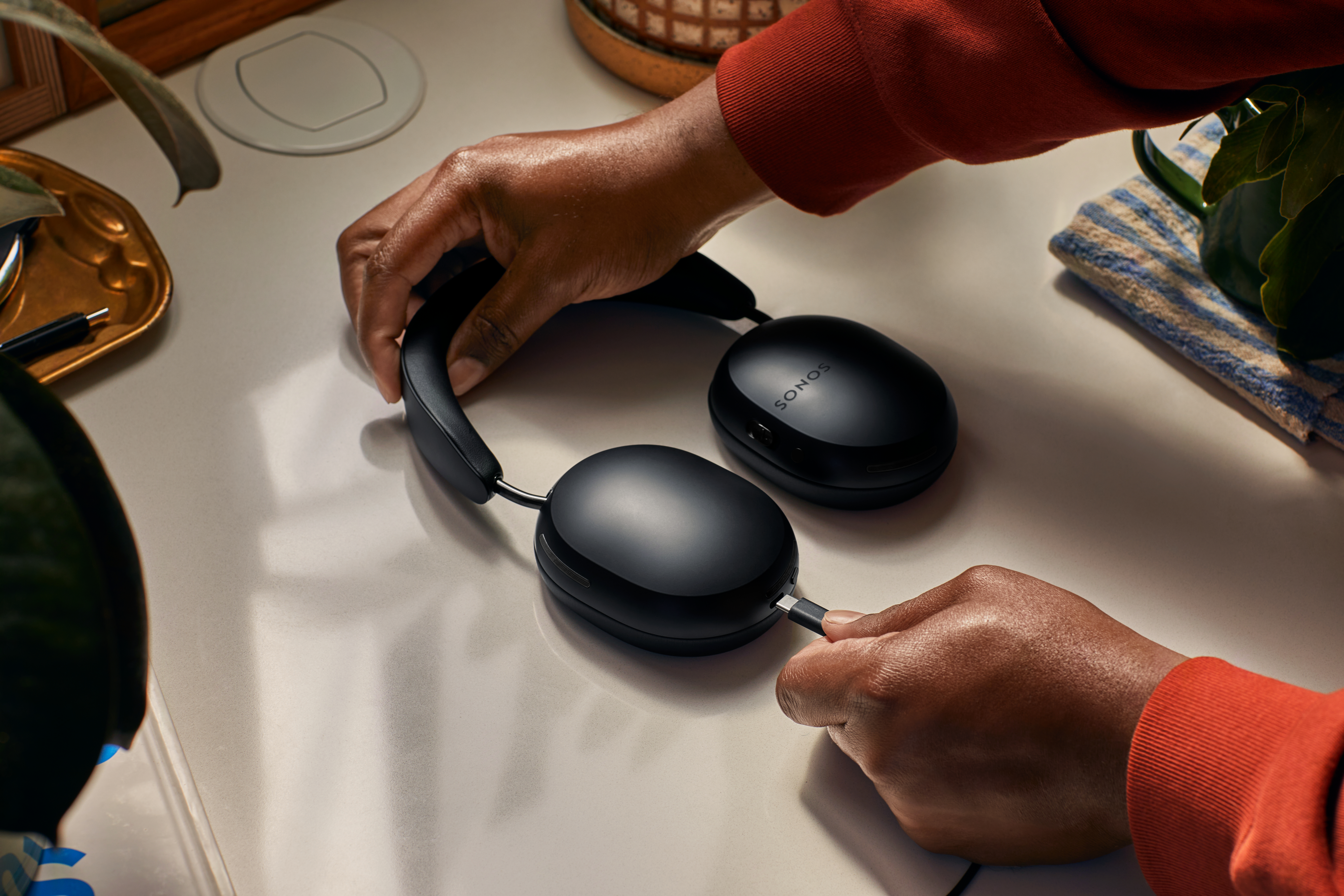 Person schließt ein schwarzes Kopfhörer-Ladekabel an. Weißer Tisch, grüne Pflanze.