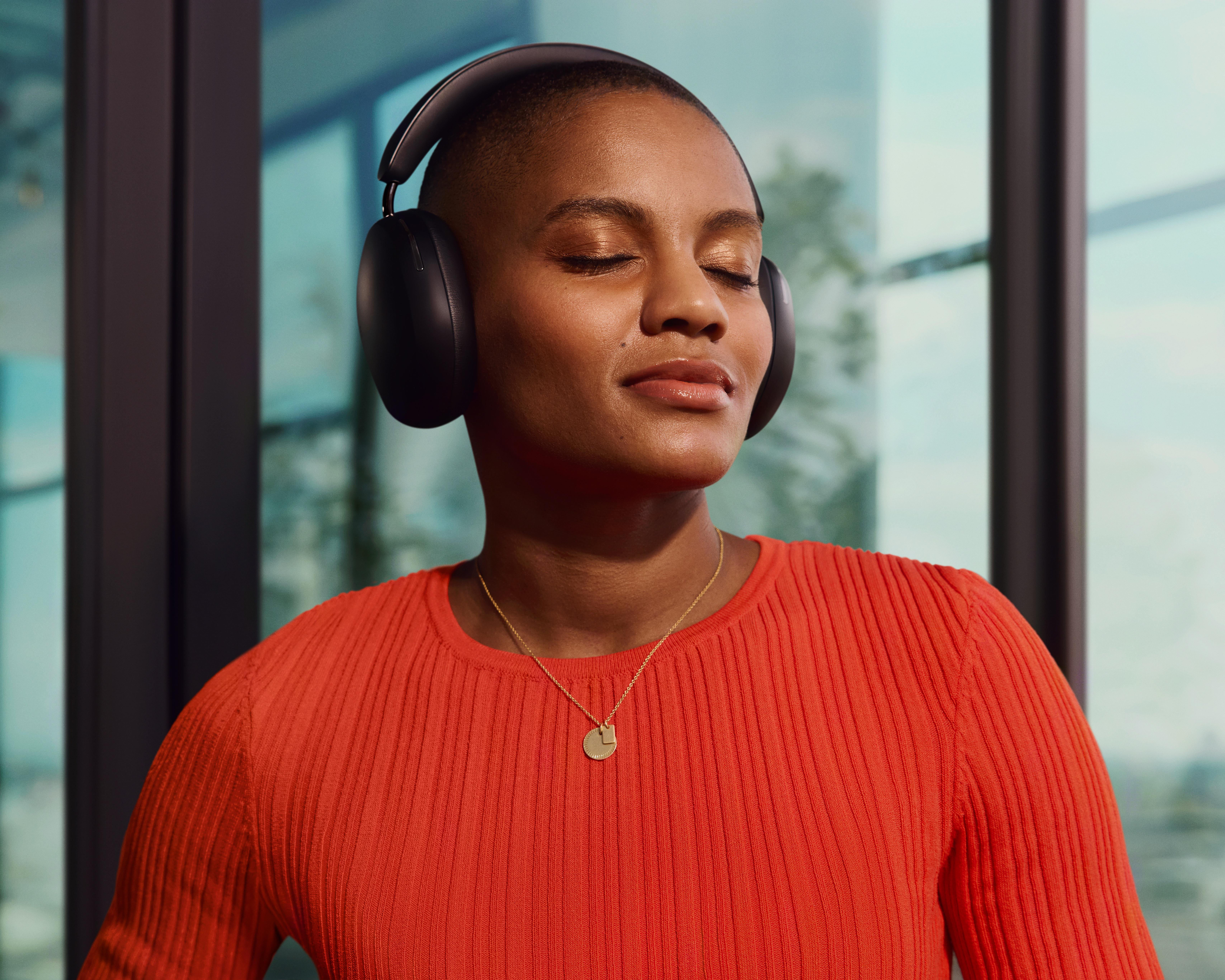 Eine Frau mit geschlossenen Augen trägt einen schwarzen Kopfhörer. Sie hat ein orangefarbenes Hemd und eine goldene Halskette.