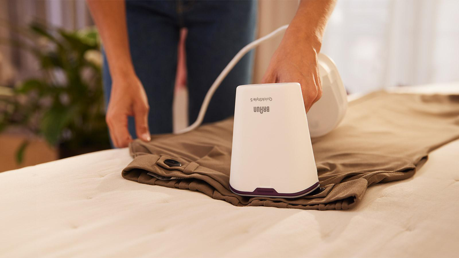 Eine Person bügelt braune Hosen mit einem weißen Braun QuickStyle 5 Bügeleisen auf einem weißen Bett.