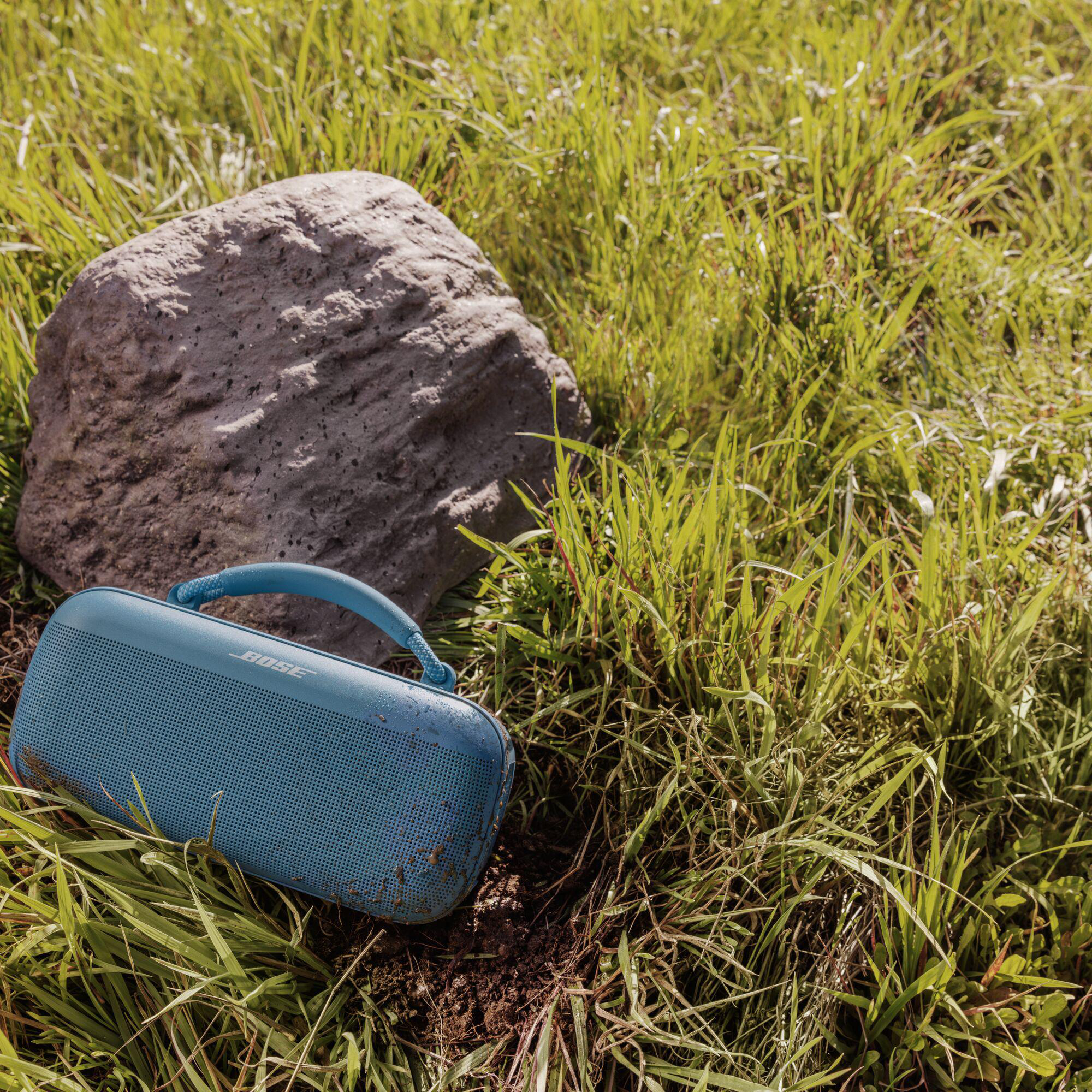 Ein blauer Bose-Lautsprecher liegt auf dem Gras neben einem großen grauen Felsen. Der Lautsprecher hat einen Griff.