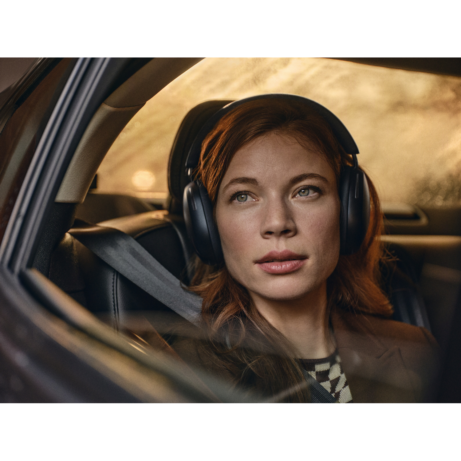 Vrouw in auto met koptelefoon. Ze heeft rood haar en kijkt vooruit. Veiligheidsgordel zichtbaar.