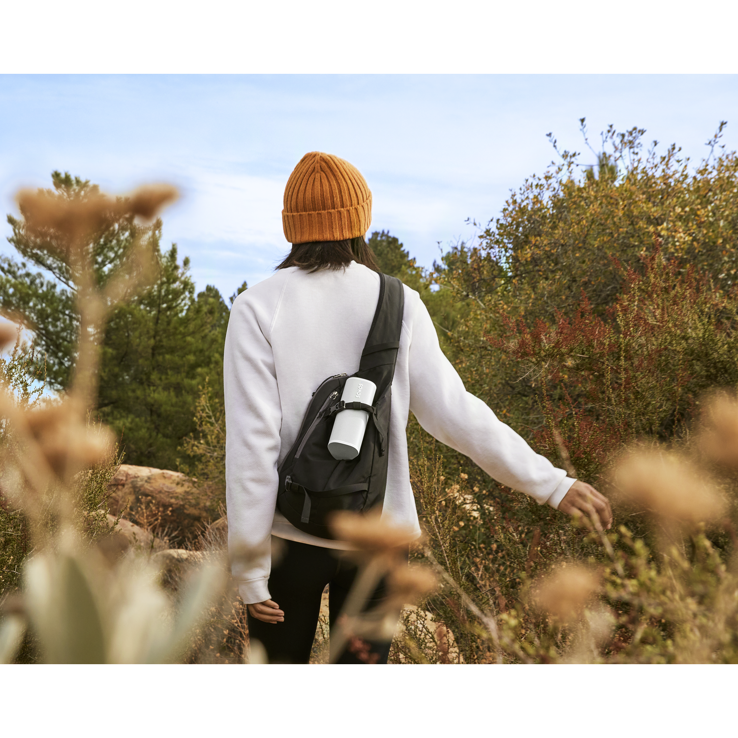 A woman in a beanie and white sweater walks through a wooded area.