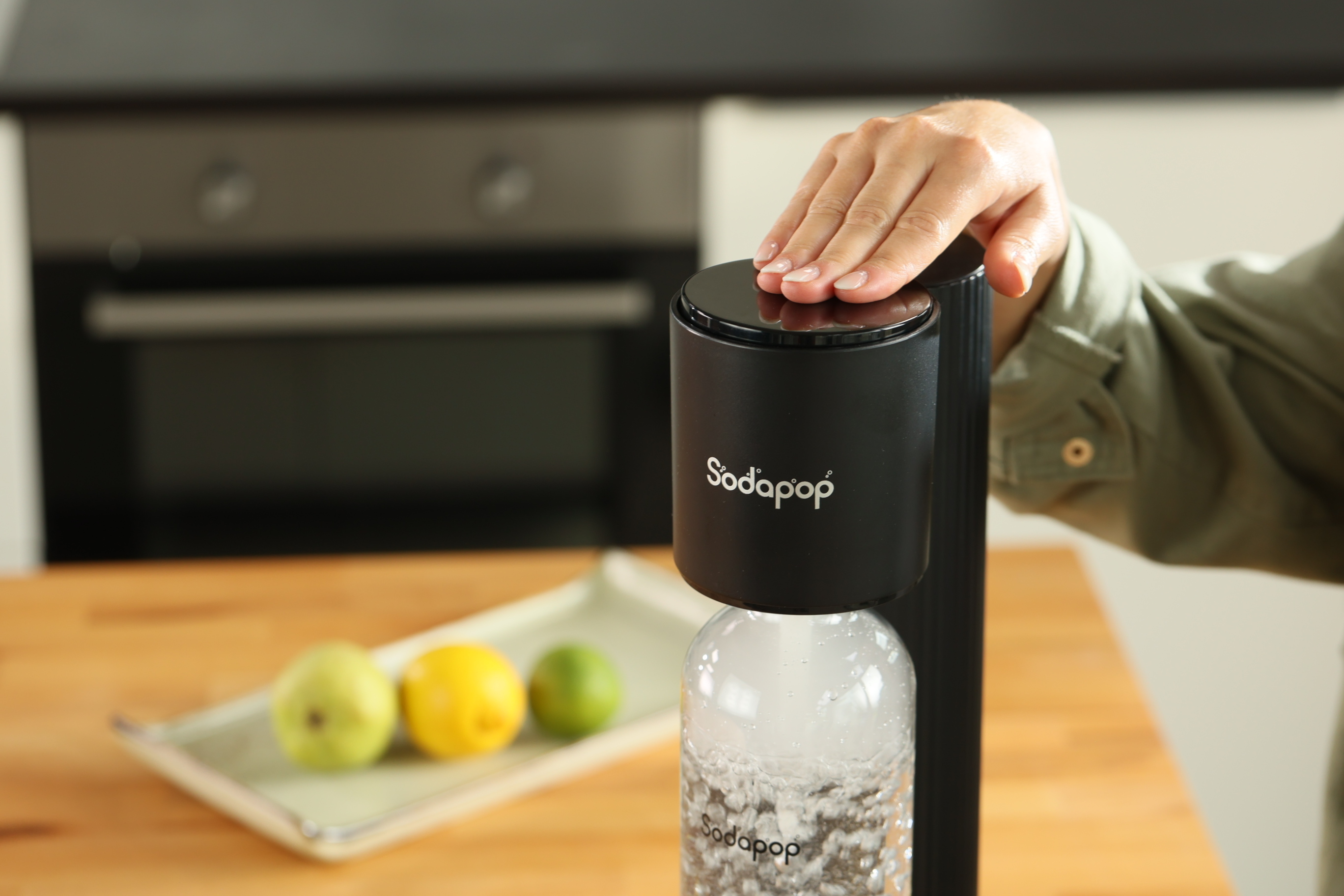 Eine Person bedient einen Soda-Maker mit einer Flasche, Früchte auf einem Tablett davor.