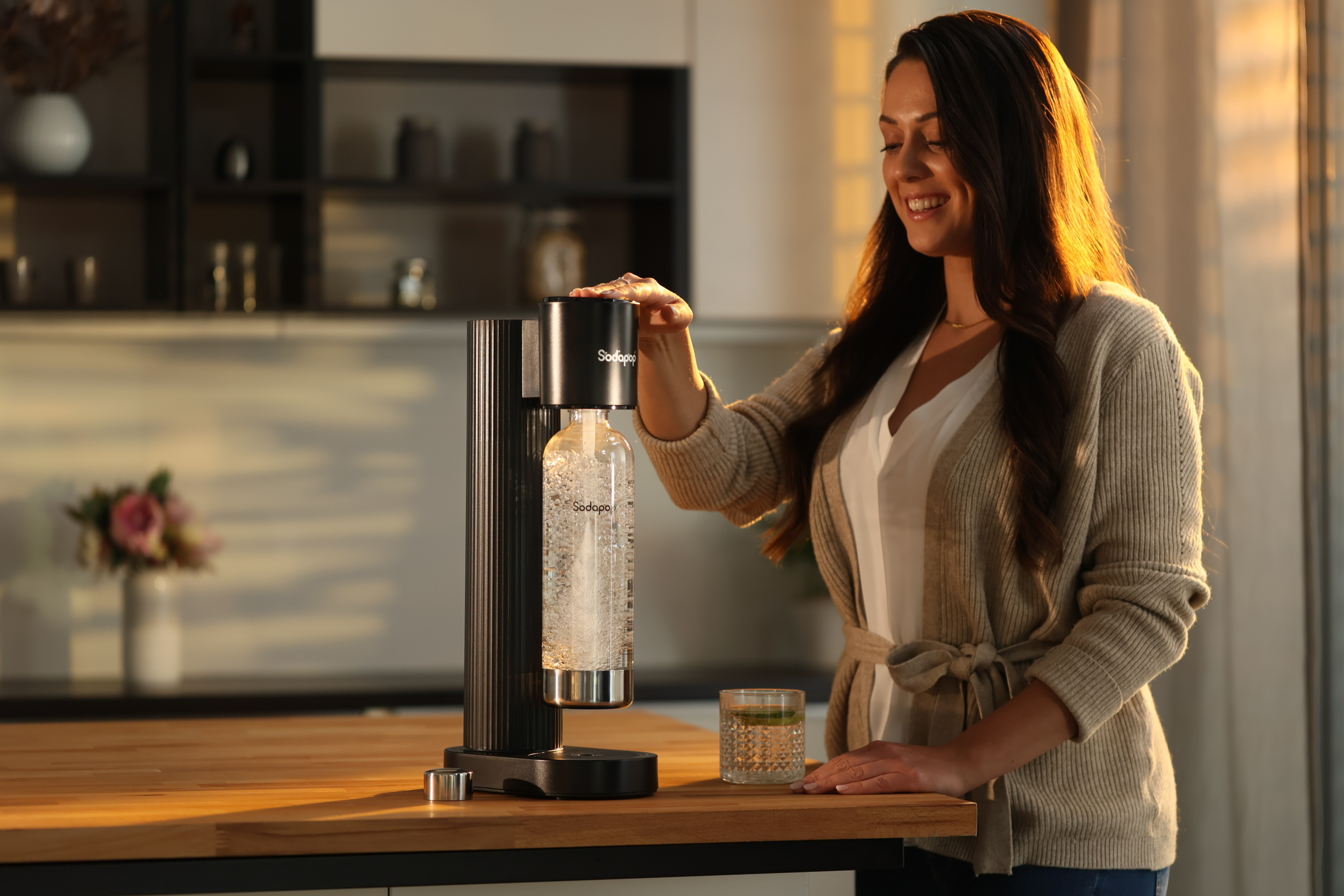 Frau macht Soda mit einem SodaStream. Sie lächelt und befindet sich in einer Küche. Ein Glas Wasser ist vorhanden.