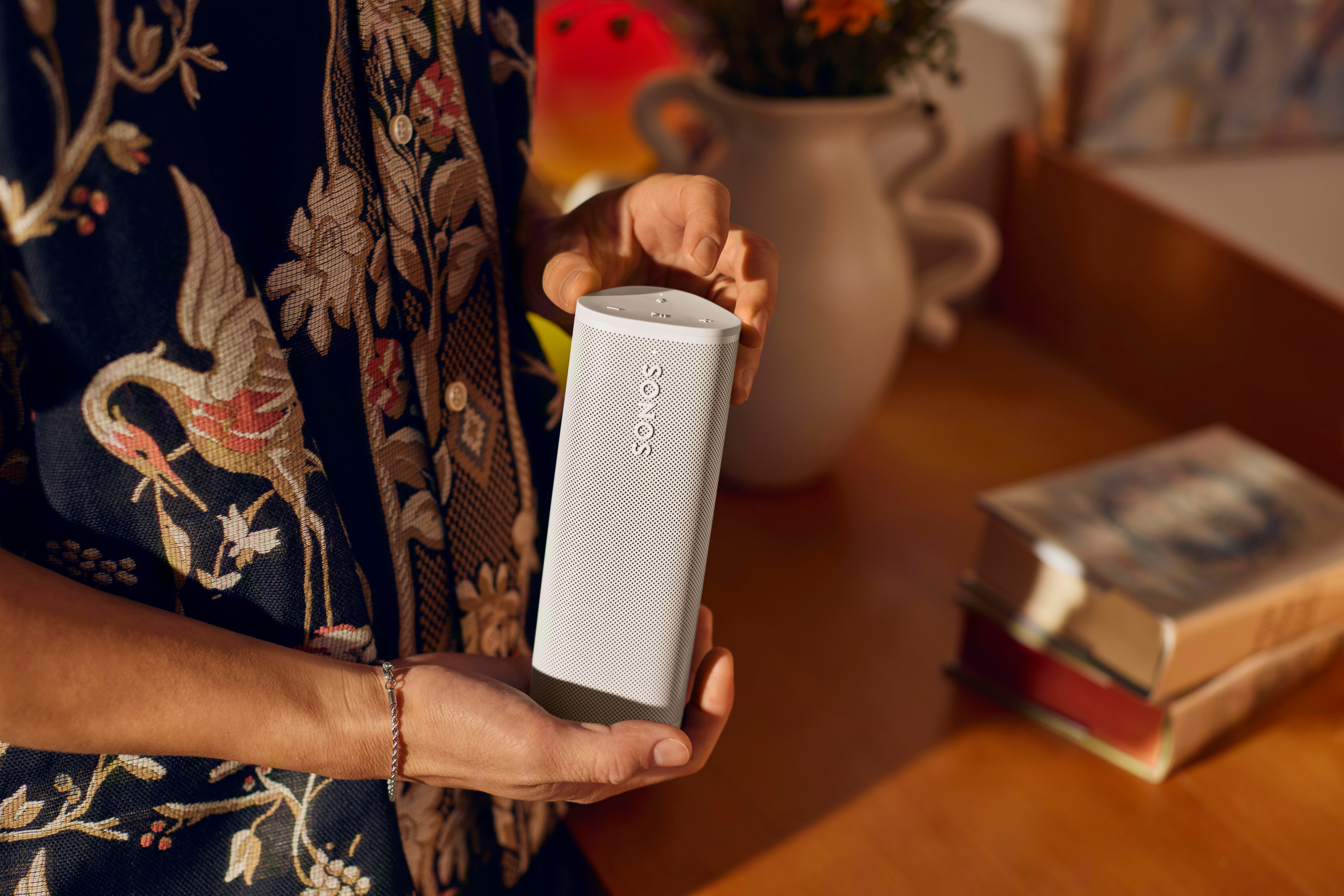 Person hält einen weißen Sonos-Lautsprecher. Hintergrund beinhaltet Bücher und Blumen.