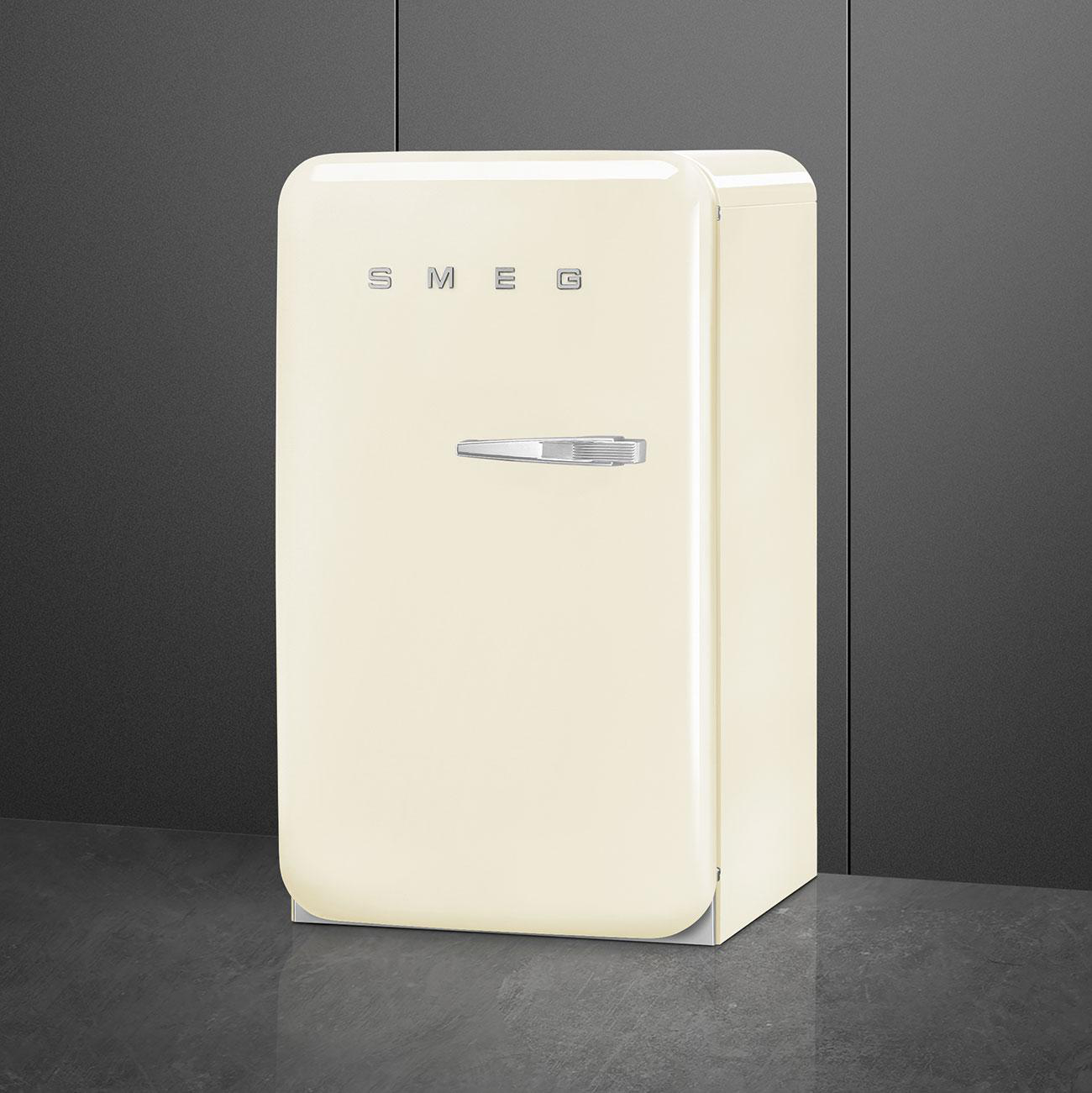 Cremefarbener Retro-Kühlschrank von SMEG. Chromgriff und -sockel. Steht vor dunkelgrauer Wand.