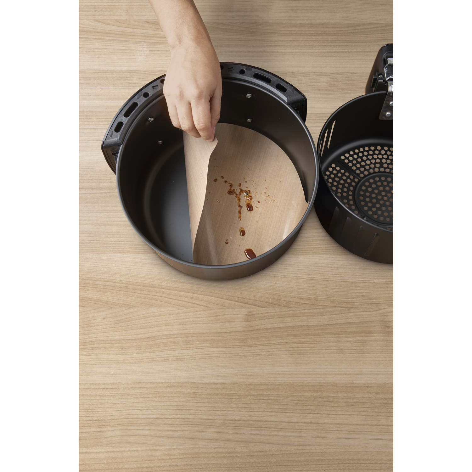 Person inserting a parchment paper into a black air fryer basket on a wooden surface.