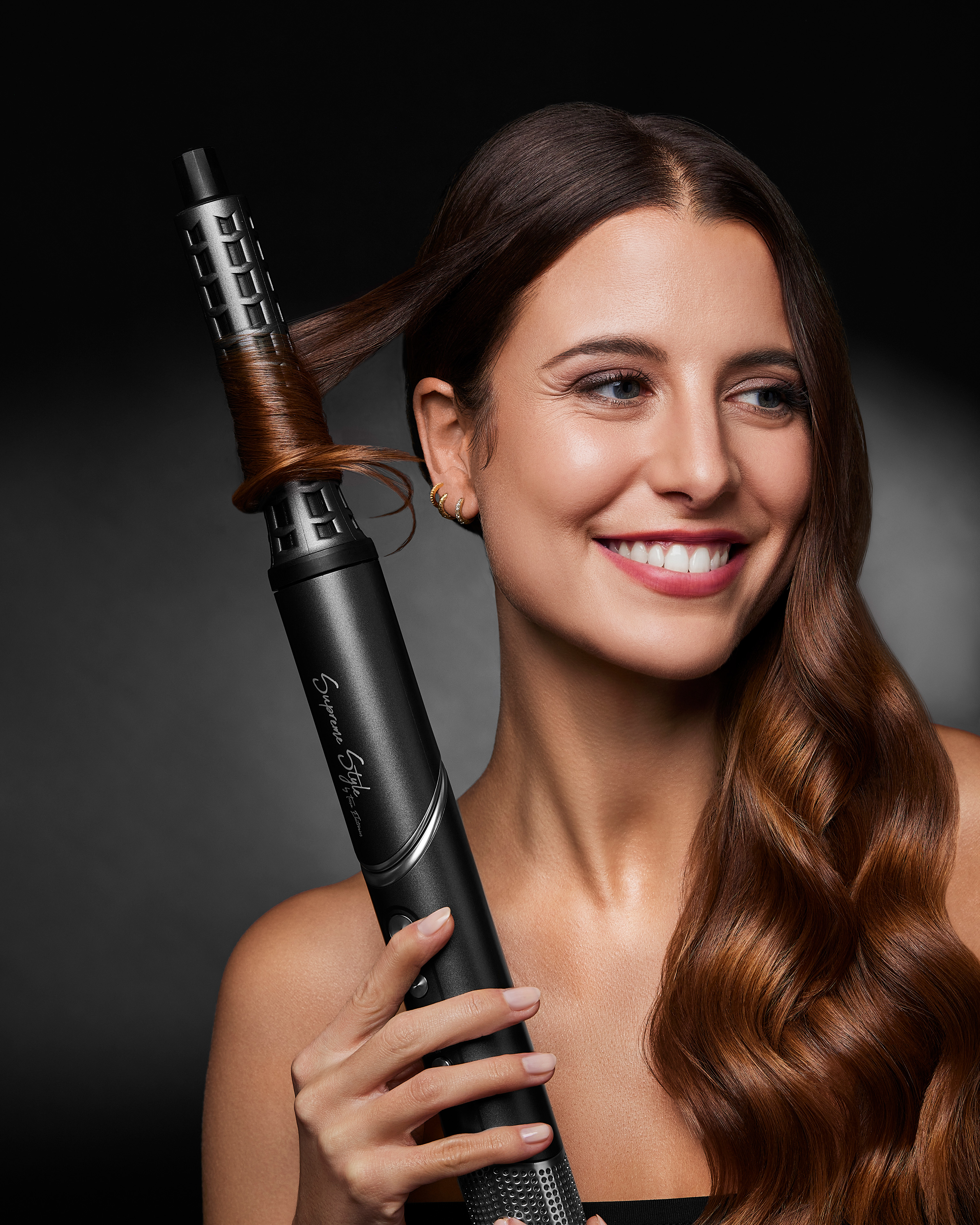 Femme avec fer à friser coiffant ses cheveux, fond noir, sourire.