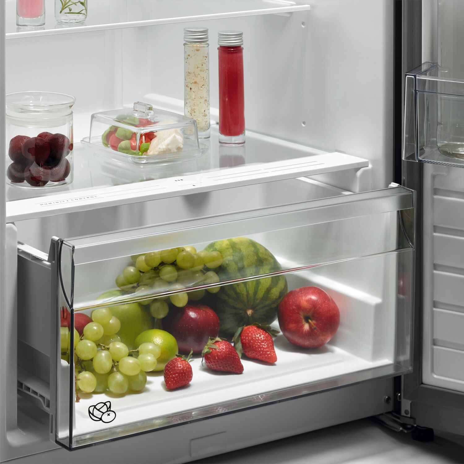 Open fridge with fruits and vegetables in a drawer, glass jars on the shelves.