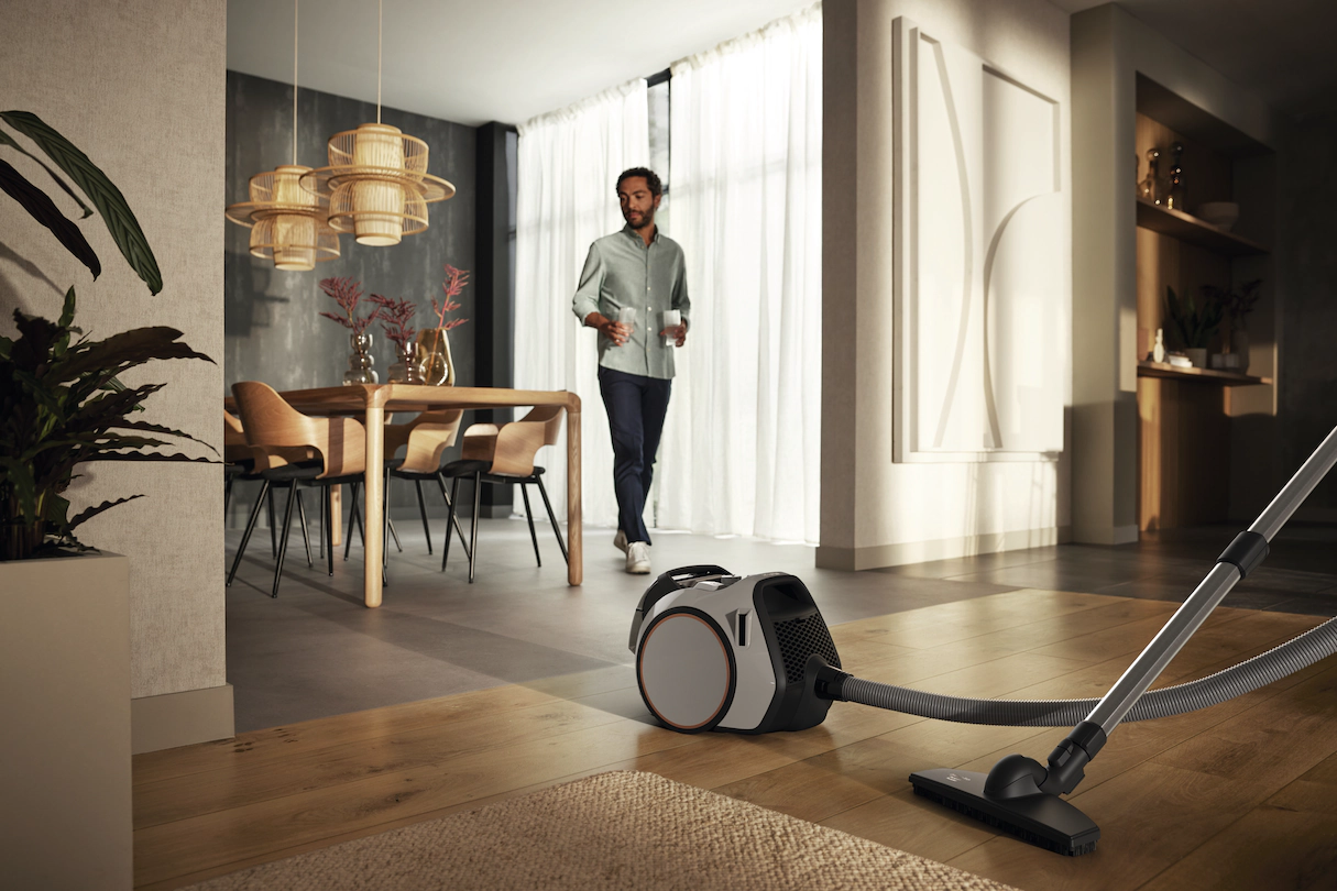 Homme marchant dans une maison moderne. Aspirateur au premier plan. Salle à manger visible en arrière-plan.