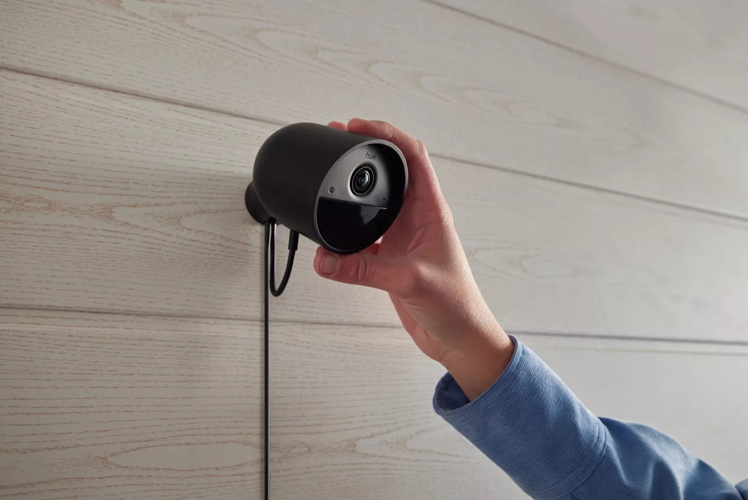 Czarna kamera bezpieczeństwa na białej ścianie z panelami drewnianymi, trzymana przez dłoń.