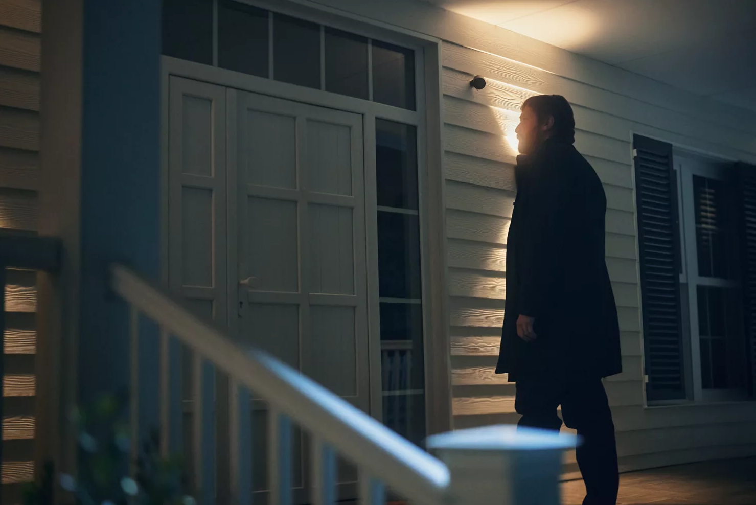 Mężczyzna w ciemnym płaszczu na werandzie w nocy. Dom z białym sidingiem i drzwiami.
