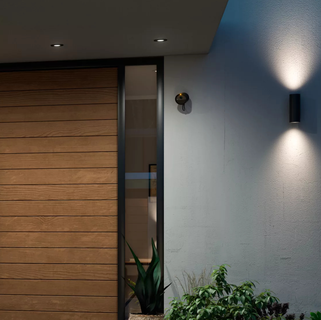 Zewnętrze domu, z drewnianymi drzwiami, oświetleniem ściennym i rośliną.