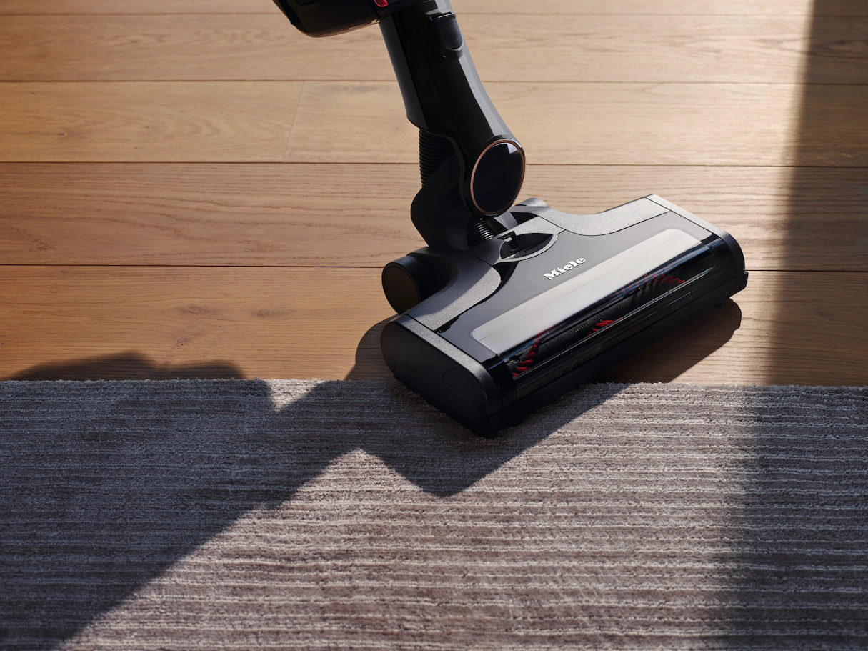 Una testina di aspirapolvere Miele nera e rossa che pulisce un tappeto grigio. Il pavimento è in legno.
