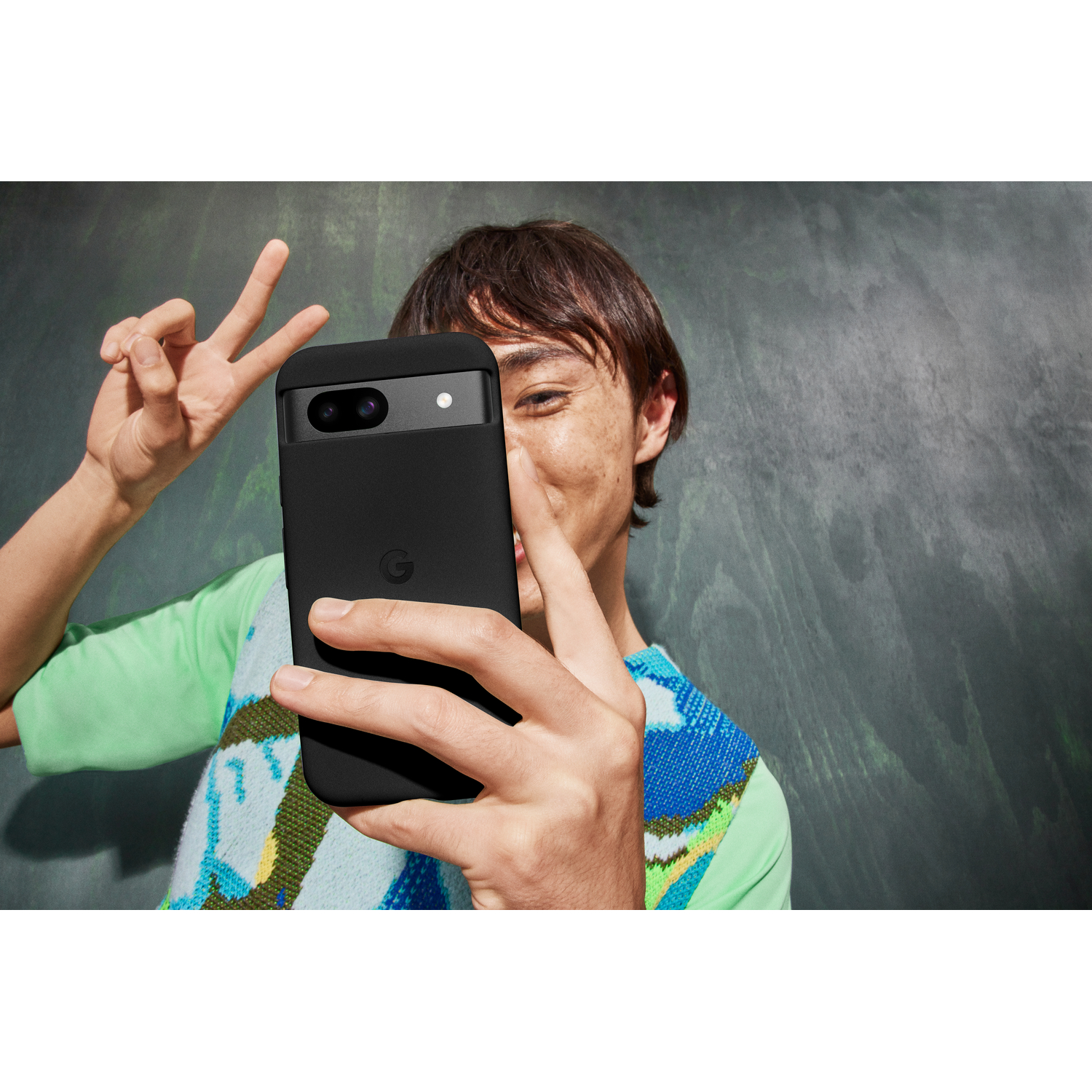 Une personne prend un selfie avec un téléphone Pixel noir dans une coque, signe de paix.