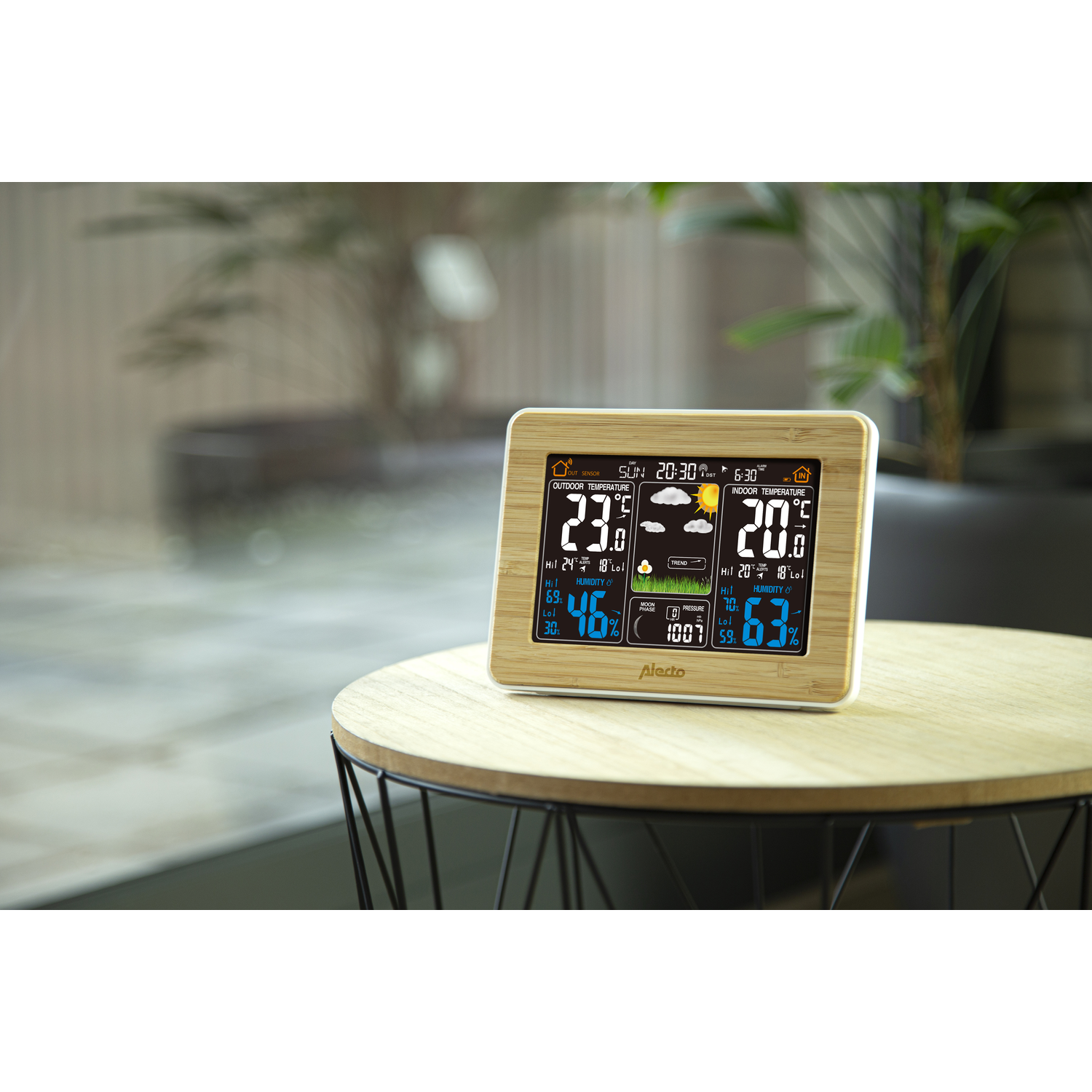 A wooden weather station on a table displays the time, temperature, and weather icons.