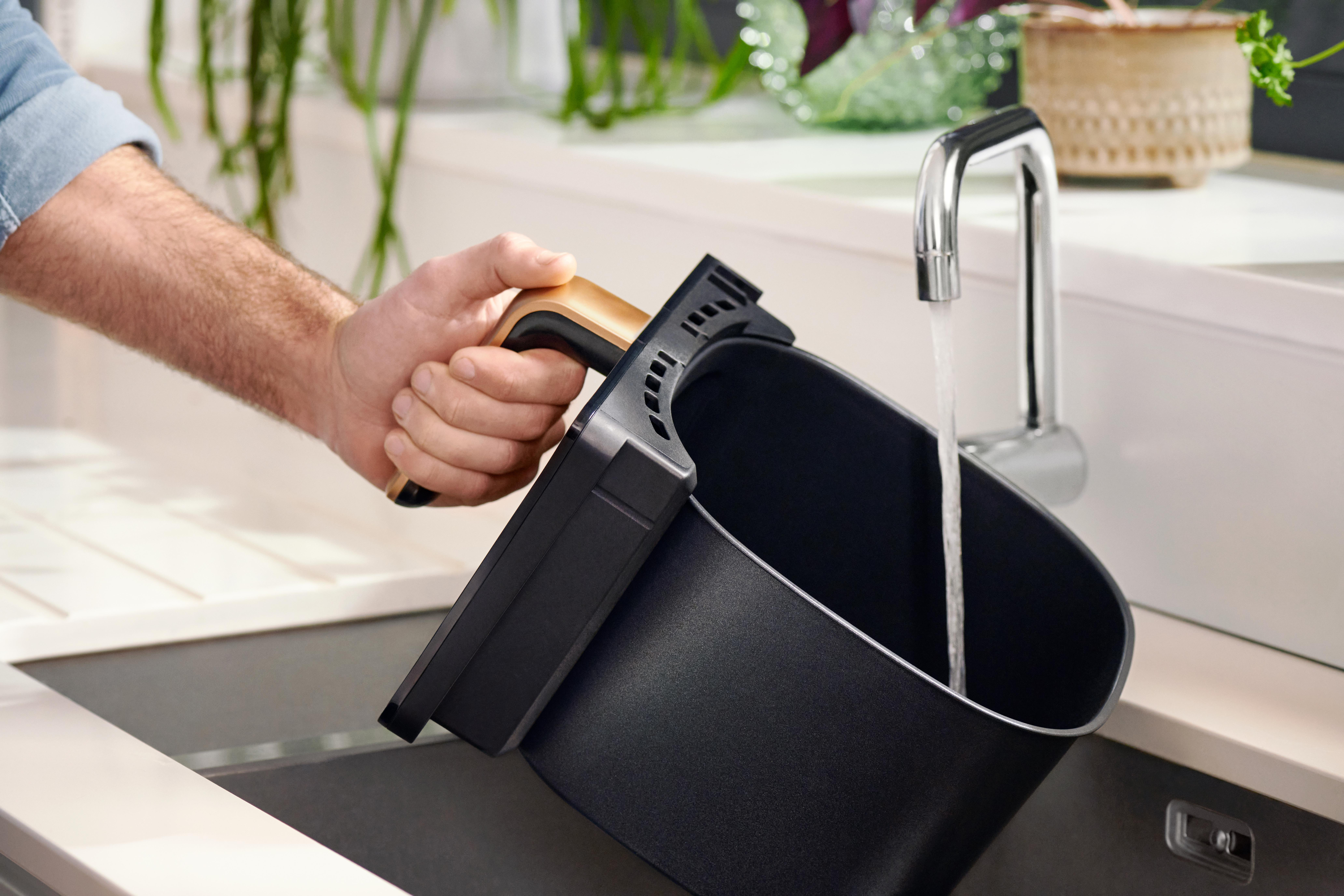 Person wäscht einen schwarzen Airfryer-Korb unter einem Wasserhahn in einem Edelstahlbecken.