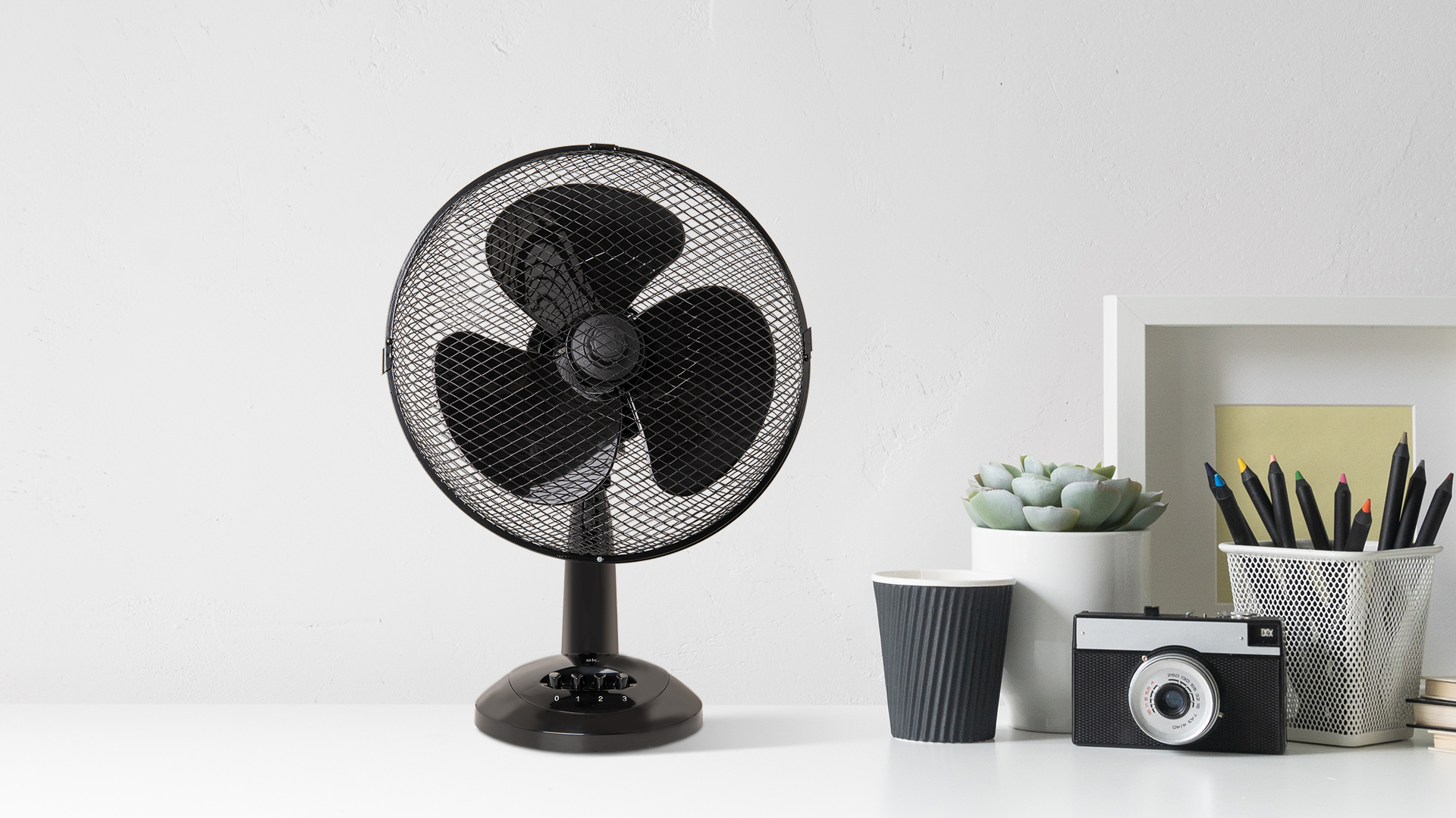 Schwarzer Tischventilator mit Schutzgitter, neben Tasse, Pflanze, Kamera und Stiften.