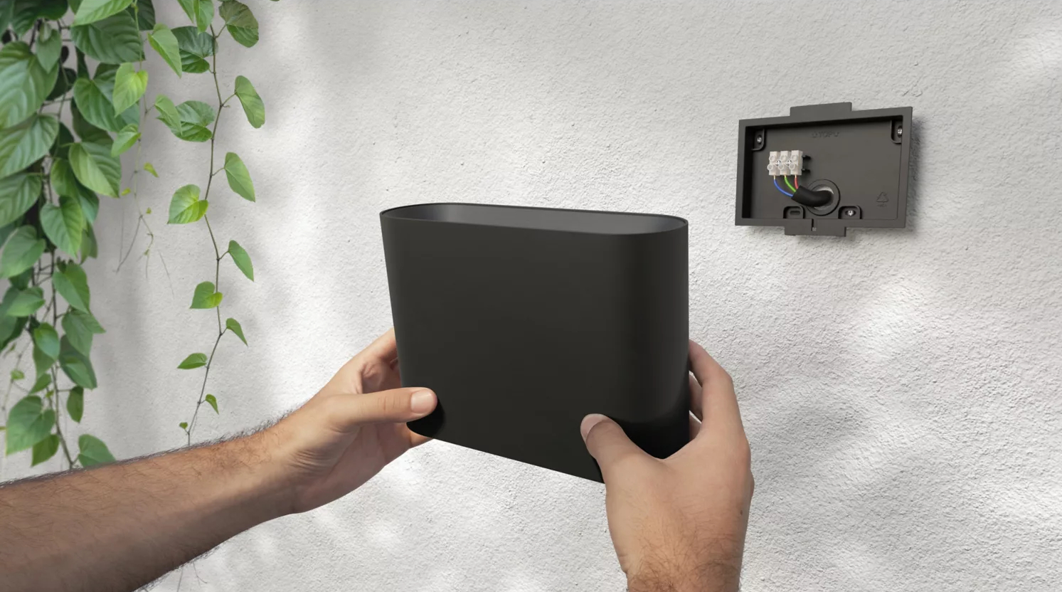 Dłonie trzymające czarną lampę ścienną, z białą ścianą i zielenią.