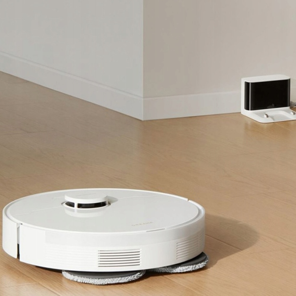 Biały robot odkurzający sprząta drewnianą podłogę. Stacja ładująca w tle.