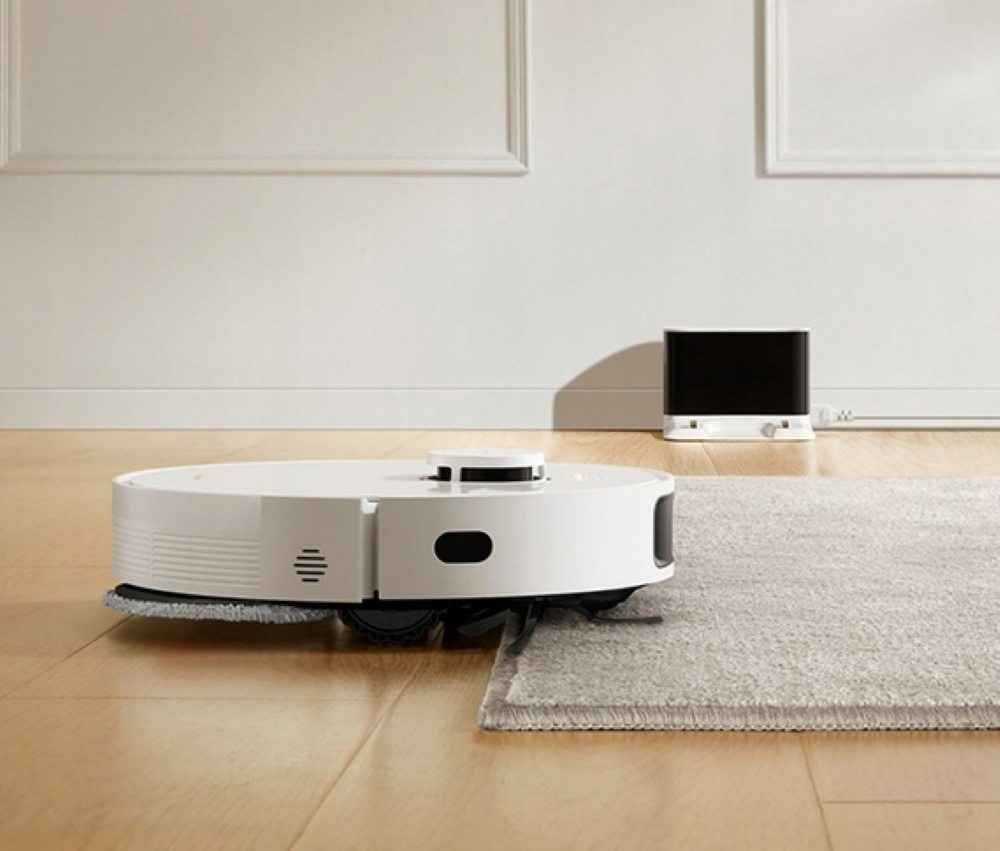 Biały odkurzacz robot sprząta drewnianą podłogę w pobliżu dywanu i ściany.