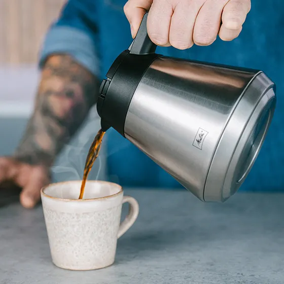 Una persona versa caffè da una caraffa d'argento in una tazza di ceramica. Vapore visibile.