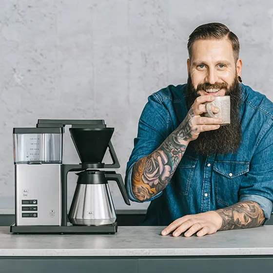Un uomo con barba e tatuaggi tiene una tazza, sorridendo davanti a una macchina da caffè.