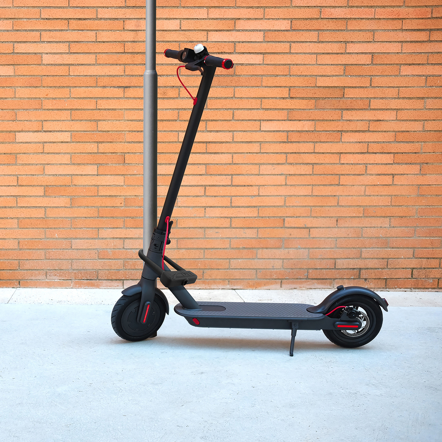 Trottinette électrique noire. Un câble rouge longe le guidon. Mur de briques en arrière-plan.