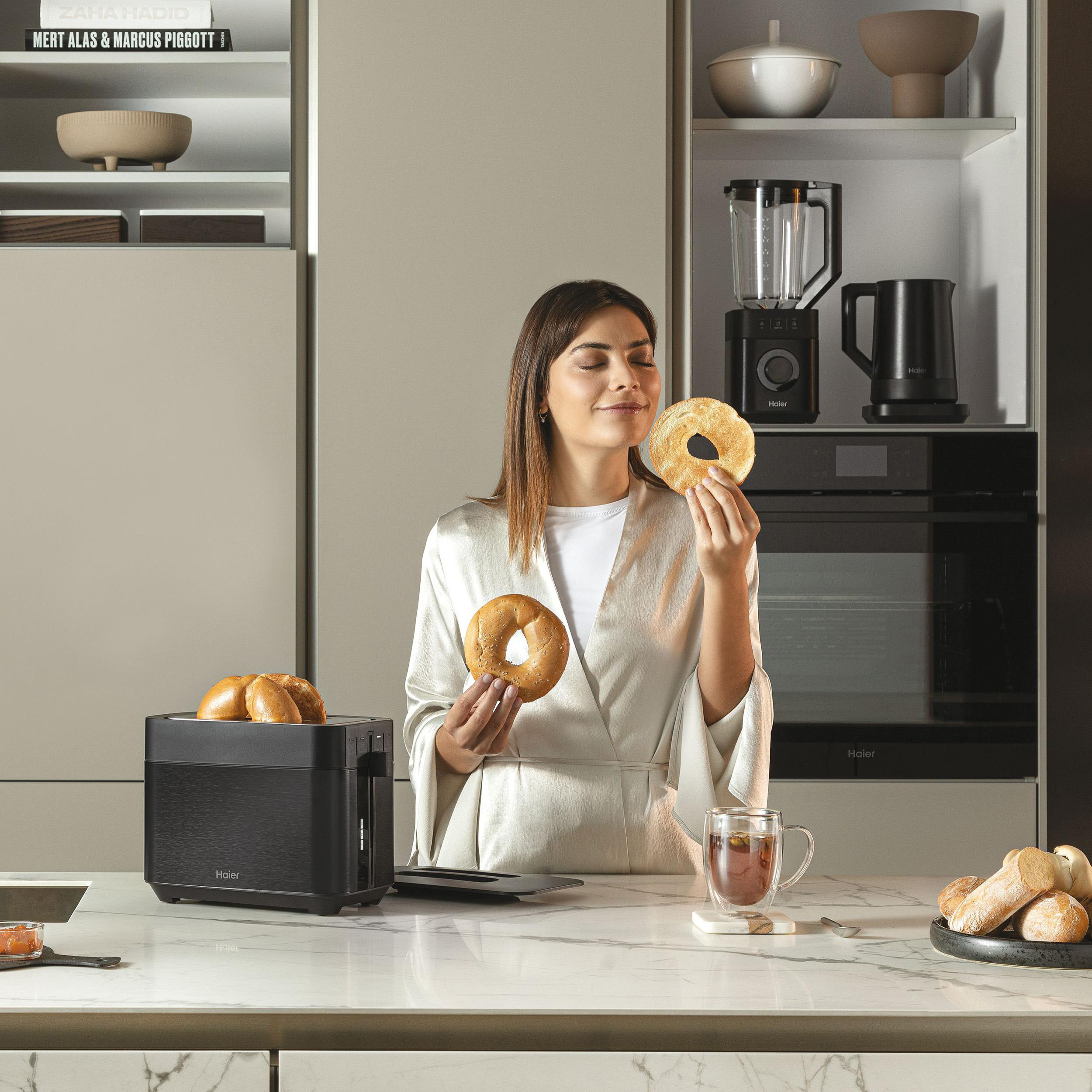 Frau hält einen Bagel, neben einem Toaster. Küche mit Mixer und anderen Gegenständen.