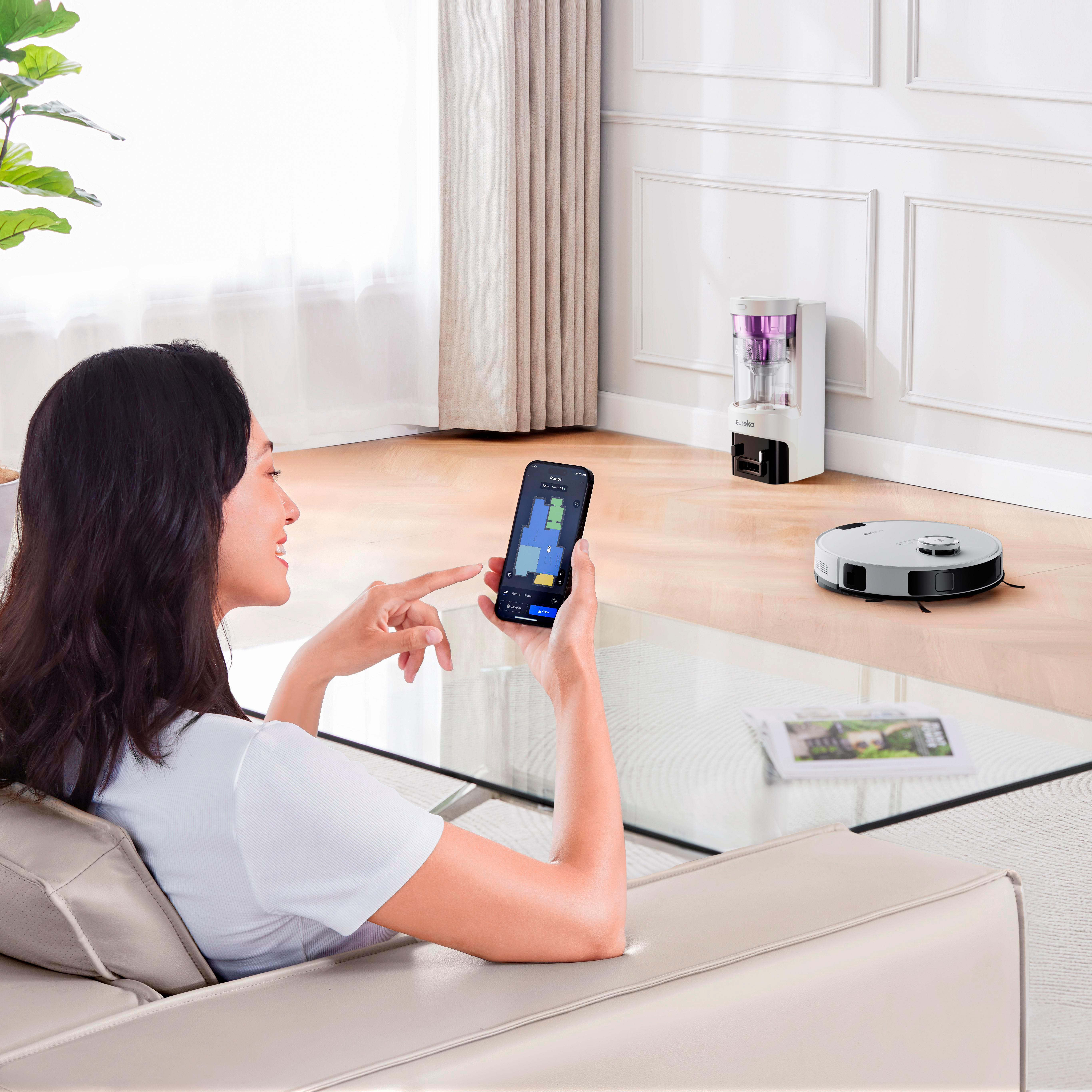 Frau benutzt ein Smartphone, um einen Roboter-Staubsauger zu steuern. Der Staubsauger ist auf dem Boden.