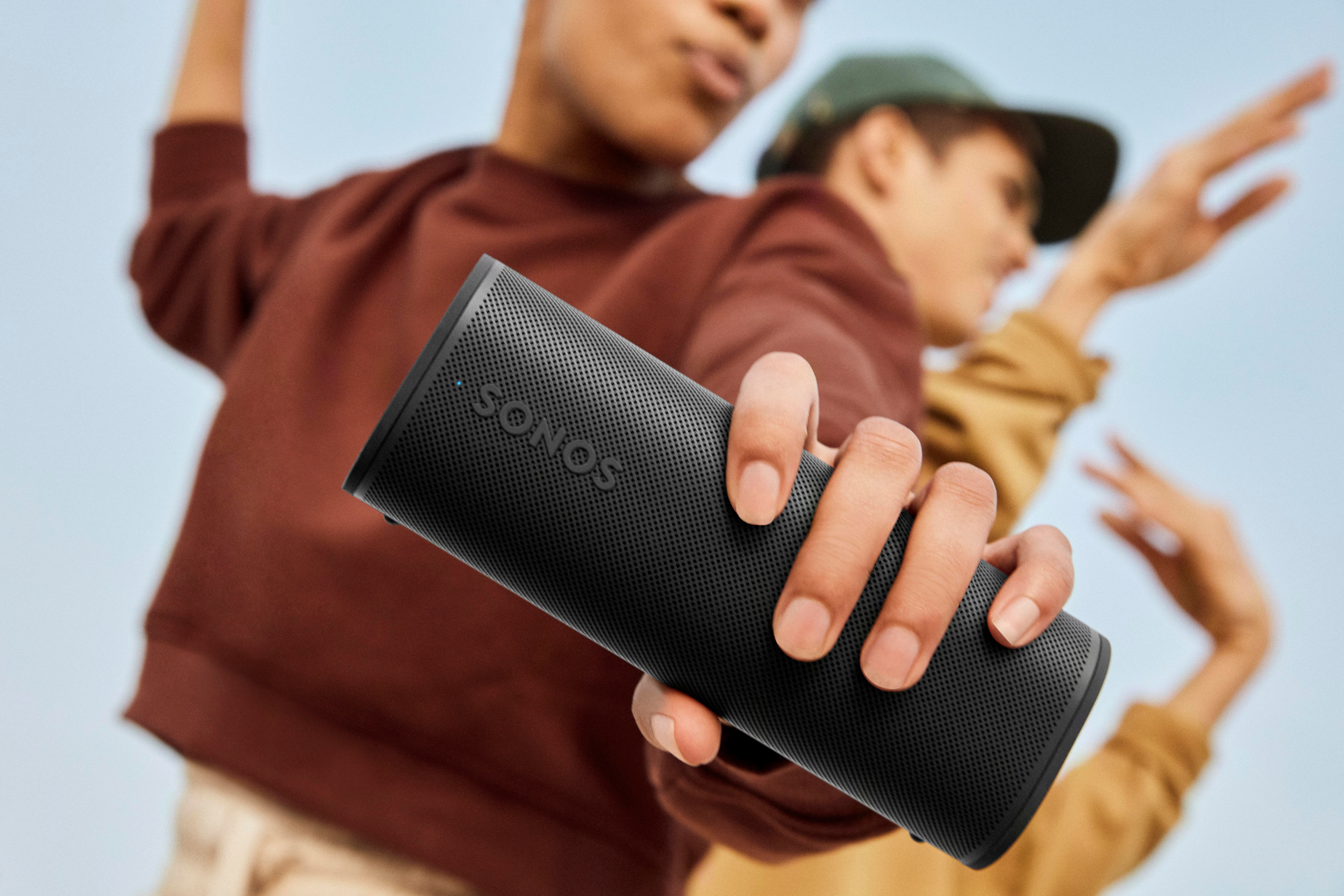 Eine Person hält einen schwarzen Sonos-Lautsprecher, im Hintergrund tanzen zwei weitere Personen.