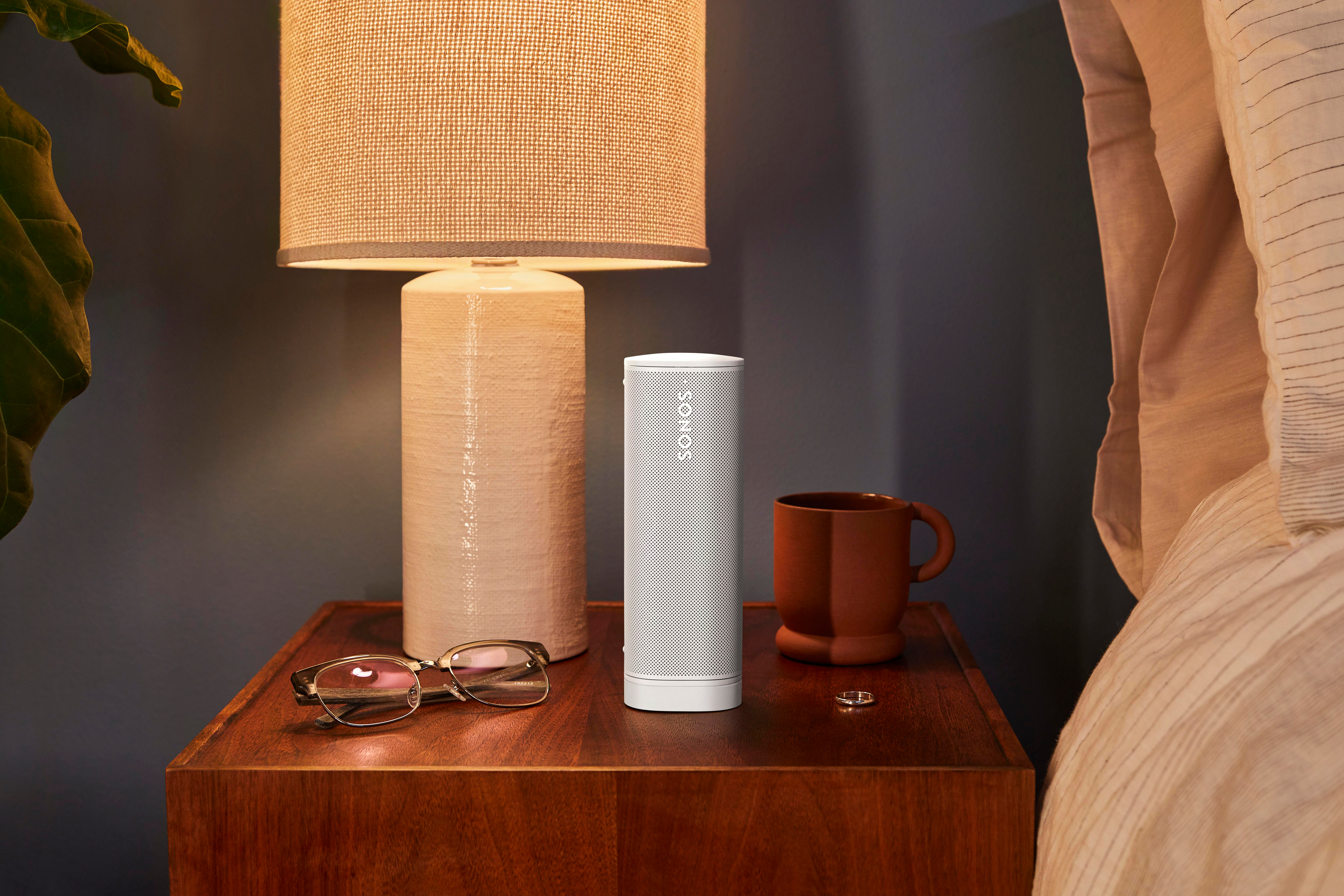 Weißer Lautsprecher auf einem hölzernen Nachttisch neben einer Lampe und einer Kaffeetasse.