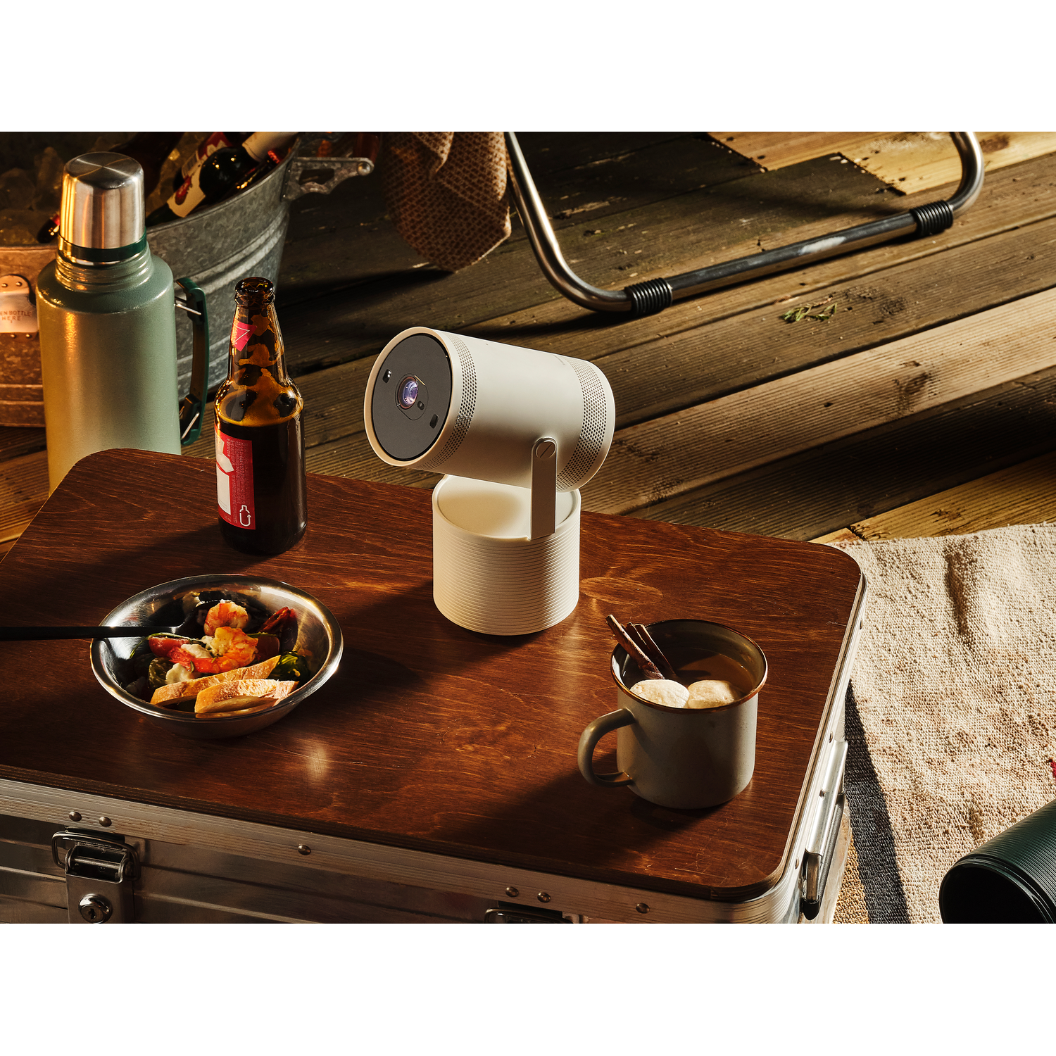 Een witte projector op een houten tafel met eten, bier en een thermoskan, buiten.