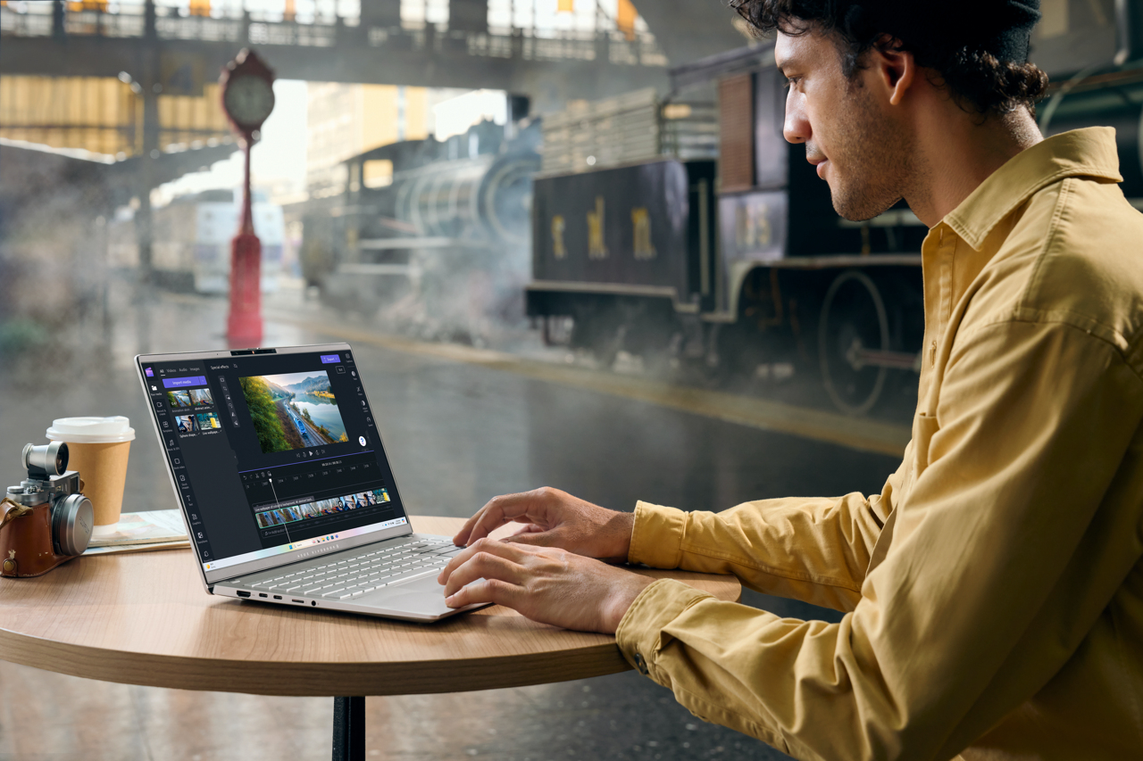Mann benutzt Laptop im Freien, bearbeitet Videos. Kaffee, Kamera auf dem Tisch. Zug im Hintergrund.